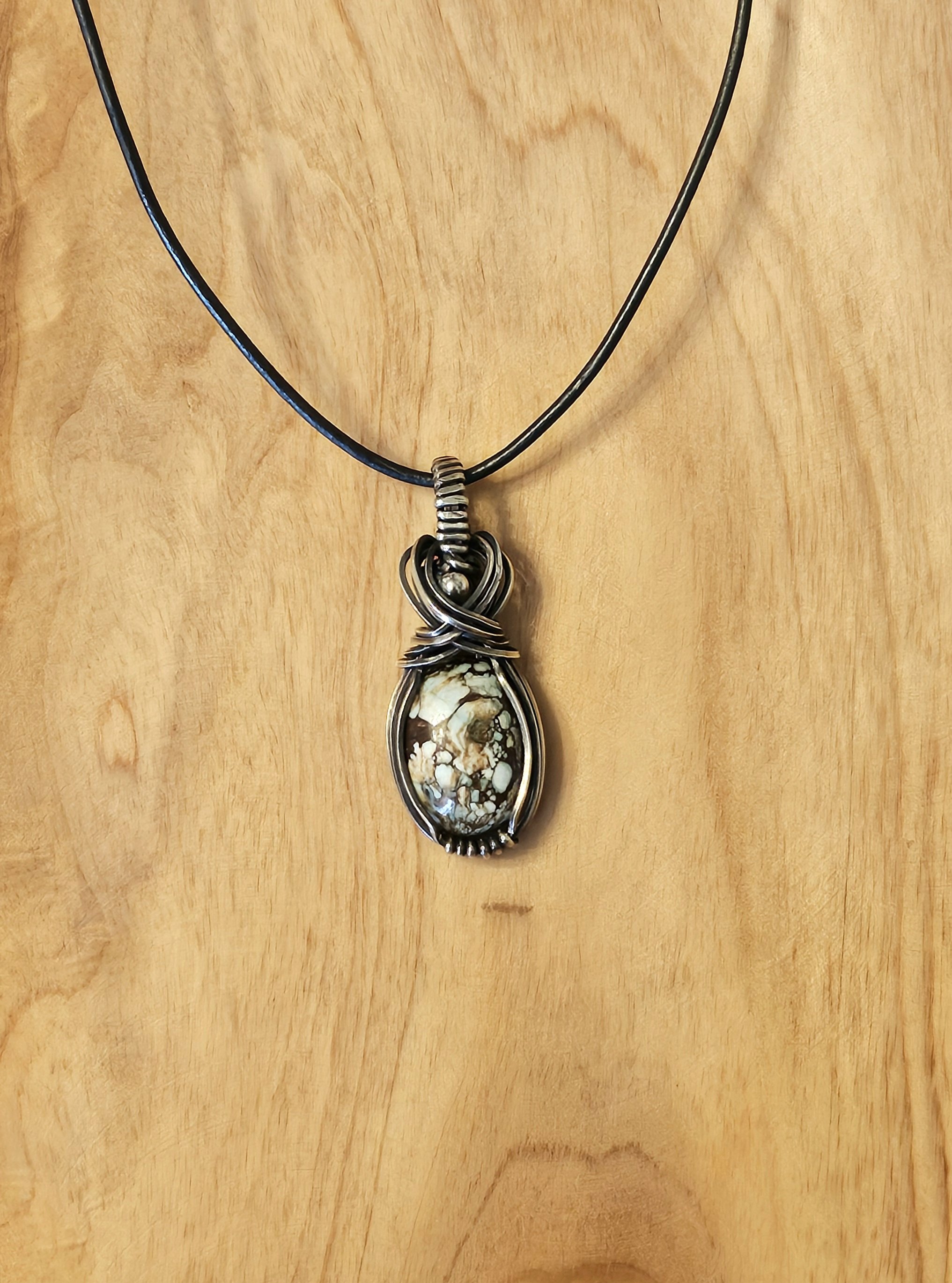Silver wire wrapped pendant with a large, multicolored stone on a black cord necklace, hanging against a light wooden background.