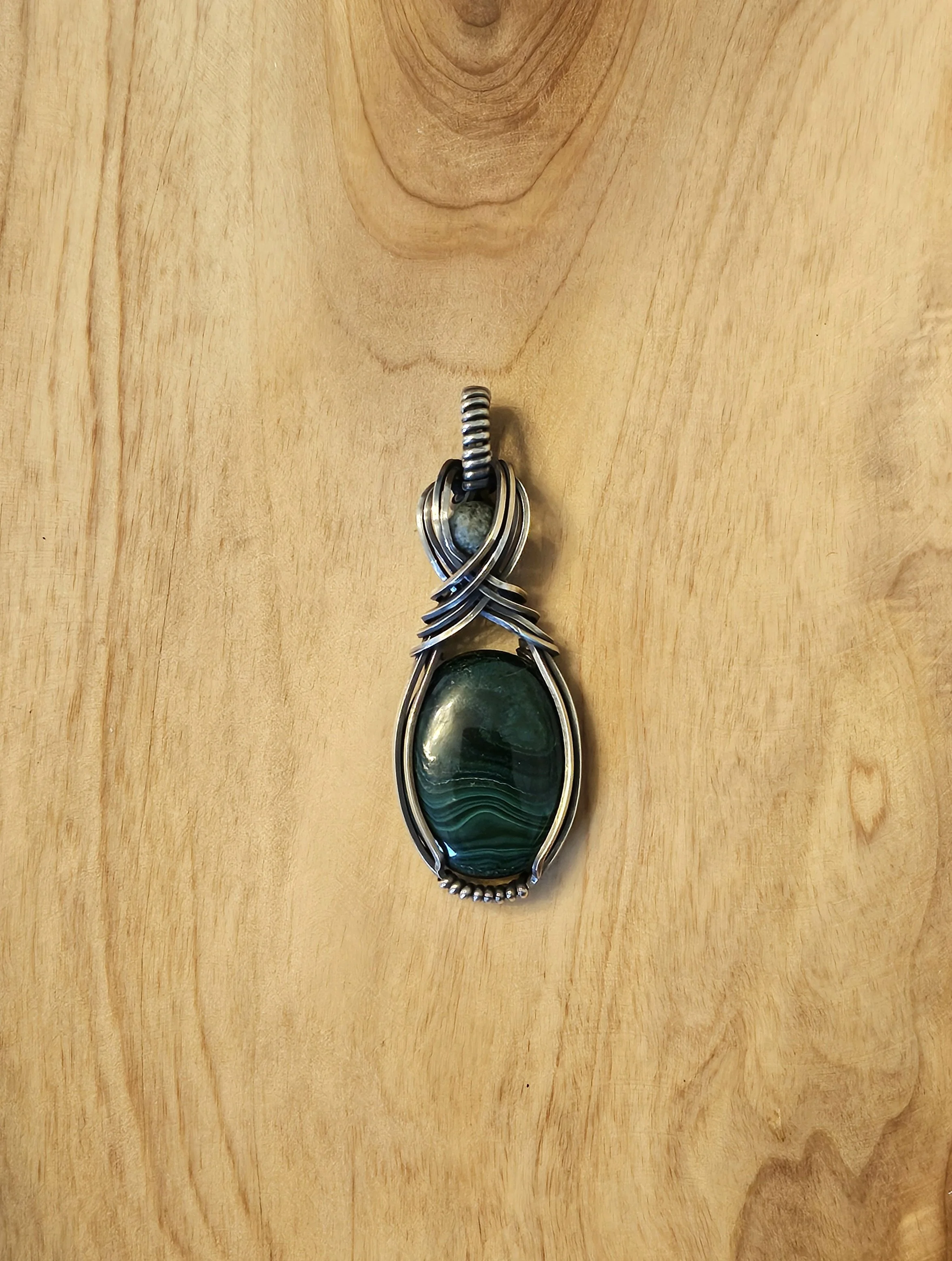 A pendant with a large green stone wrapped in silver wire, hanging from a small wire loop, placed on a wooden surface.