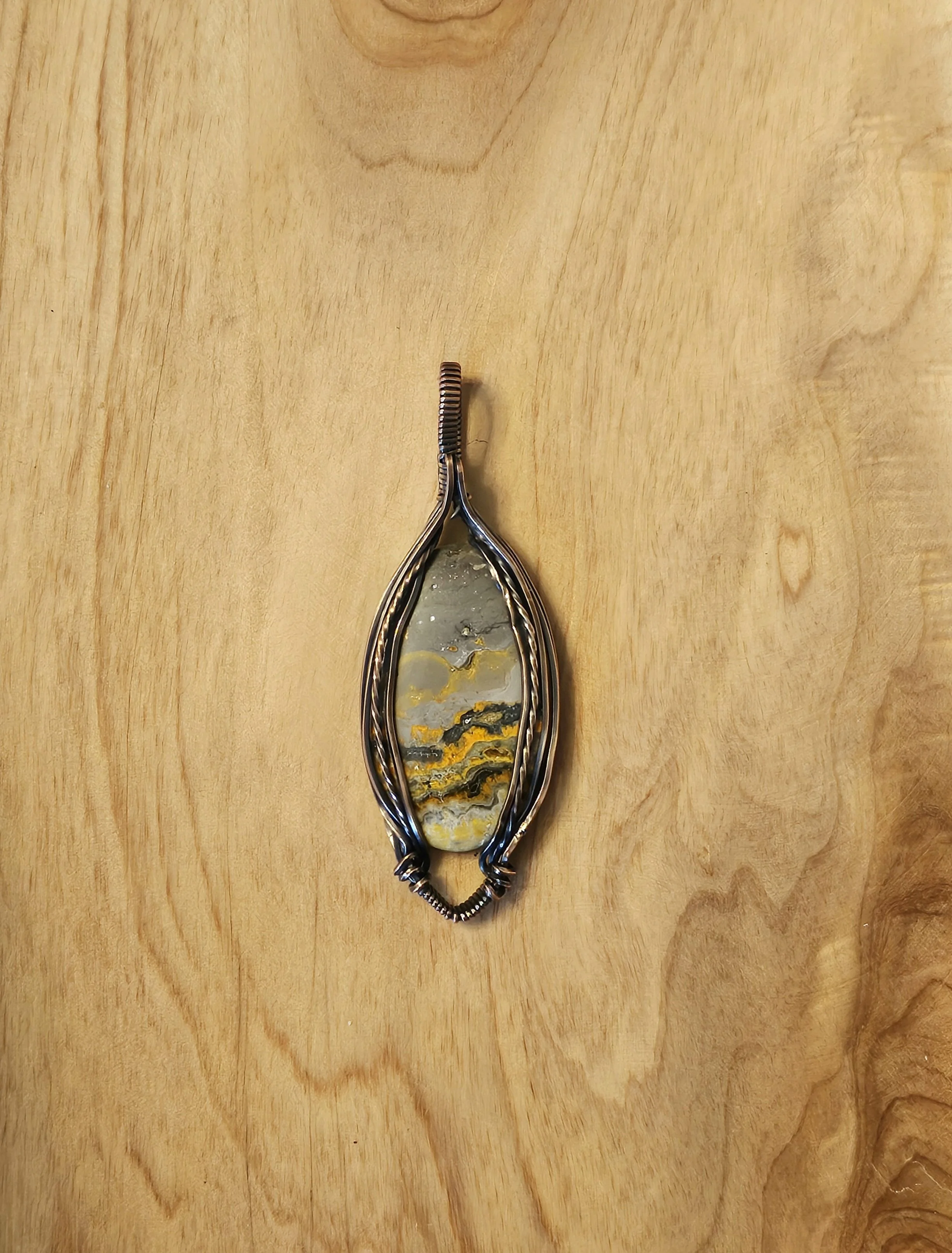 A silver pendant with a large oval stone featuring yellow, black, and gray streaks, set against a wooden background.