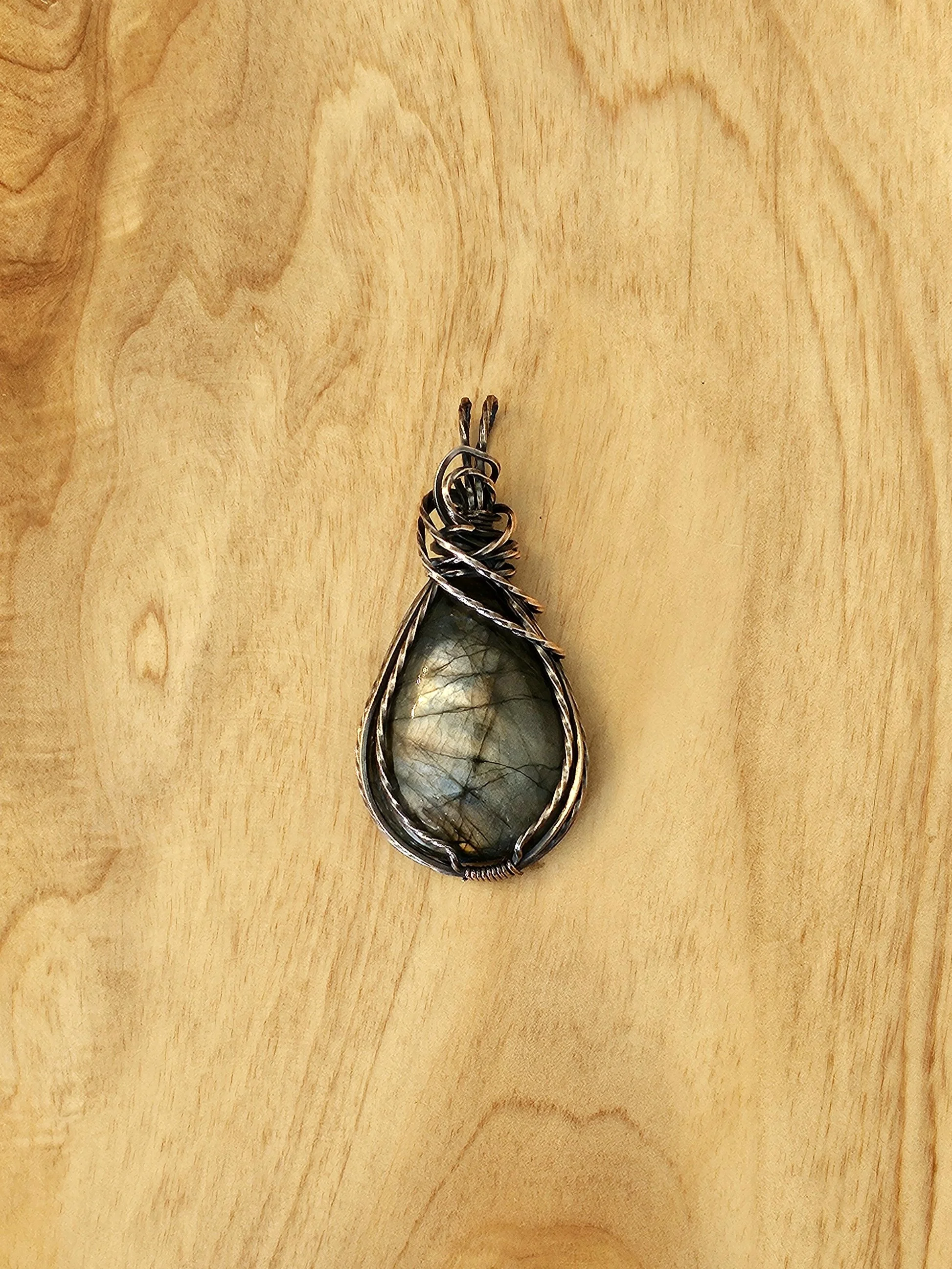 A silver wire-wrapped pendant with a large, teardrop-shaped labradorite gemstone on a wooden surface.