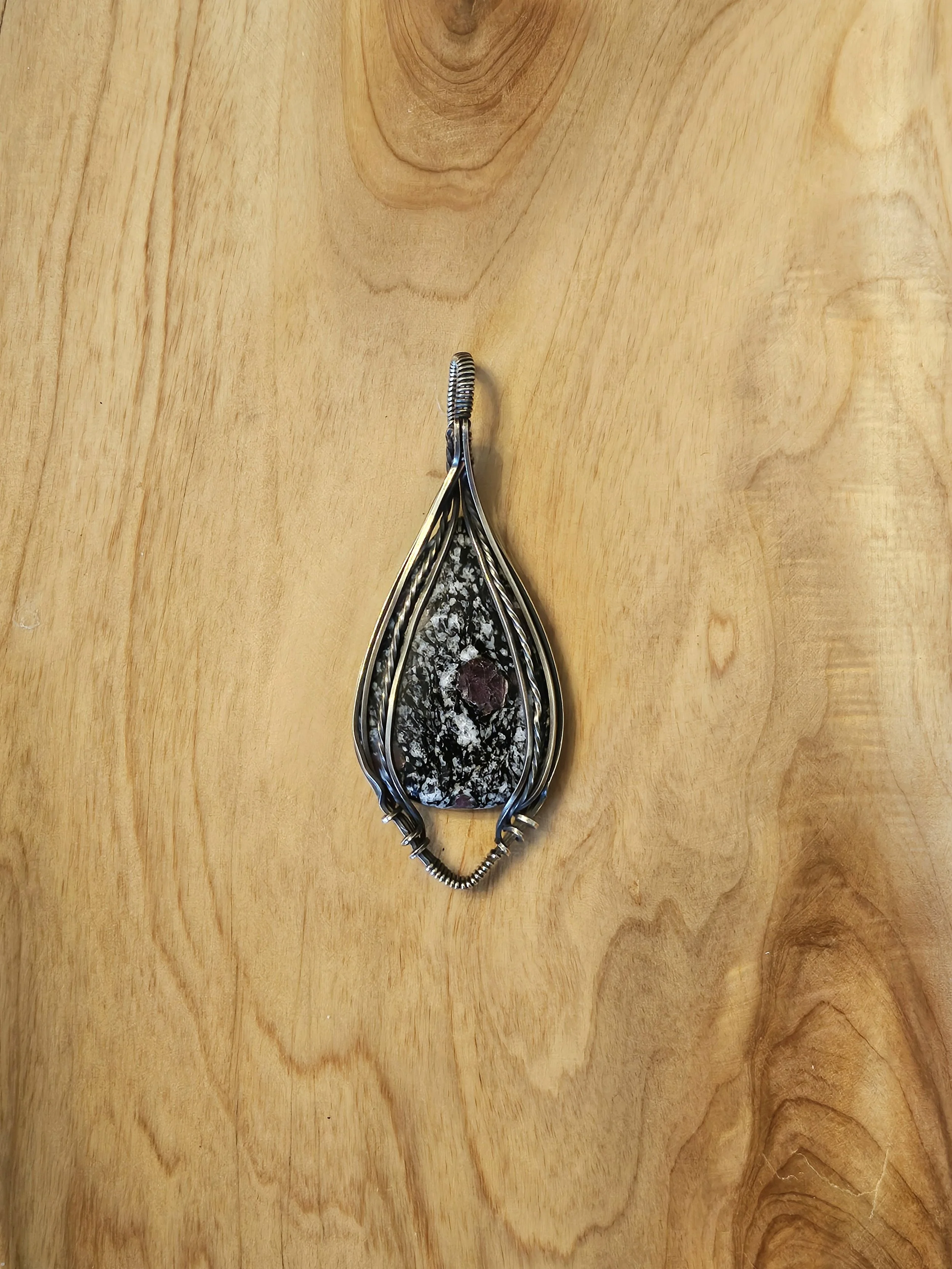 A pendant with a black and white speckled stone set in a decorative silver setting, lying on a wooden surface.