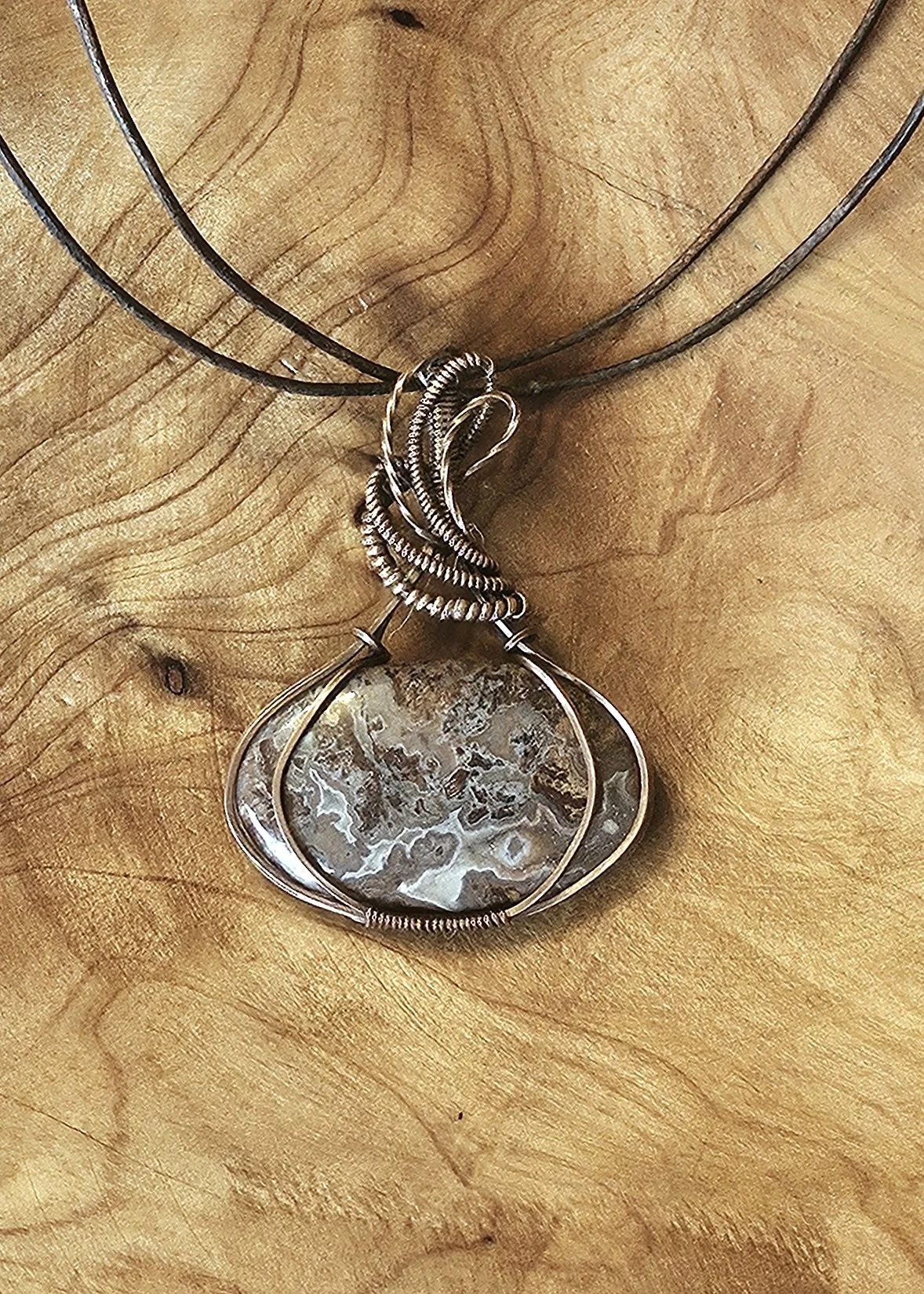 A close-up view of a necklace with a large, oval-shaped gemstone pendant featuring a banded, translucent appearance, hanging on a black cord against a wooden background.