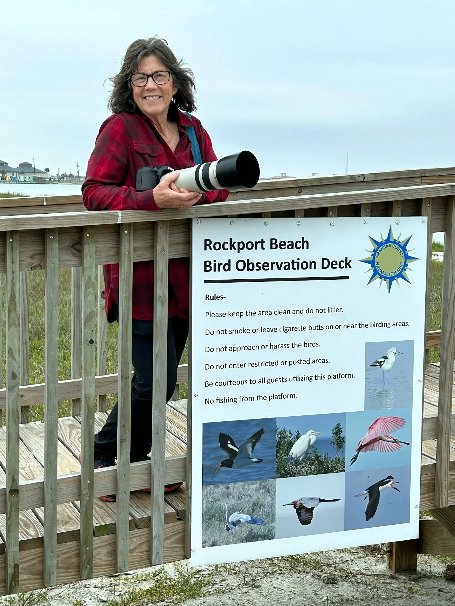 A woman with gray hair, glasses, dressed in a red plaid shirt, standing behind a wooden railing at Rockport Beach Bird Observation Deck, holding a camera with a large telephoto lens. A sign on the railing lists rules for bird watching and features images of various birds.