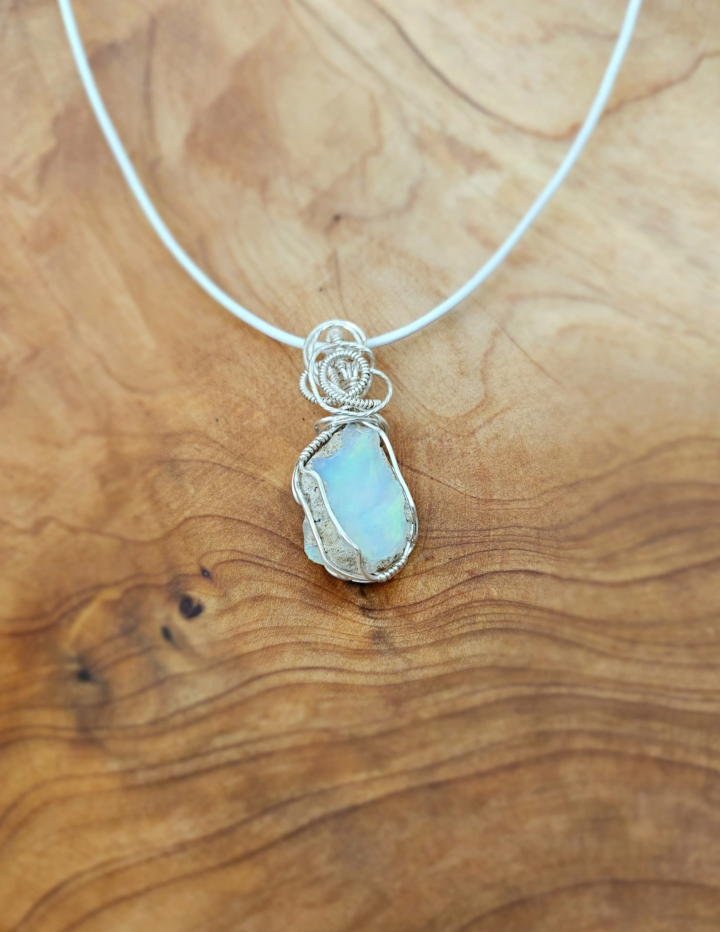 Silver wire wrapped rectangular opal pendant on a white cord necklace, resting on a wooden surface.