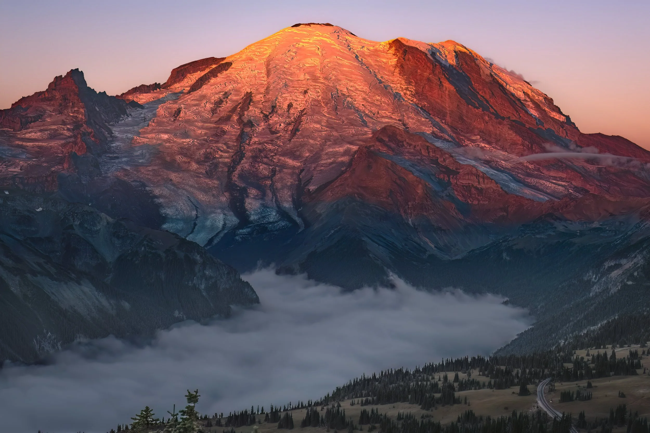 Sunset illuminating Mount Rainier with snow-capped peak, surrounded by clouds and forested valleys.