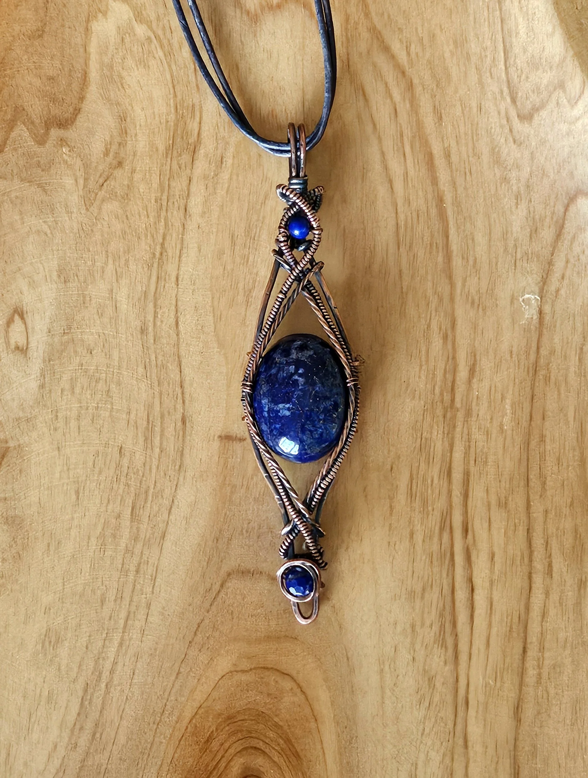 Handcrafted pendant with a large blue oval gemstone, decorated with wire wrapping, and smaller blue beads, hanging from a black cord on a wooden surface.