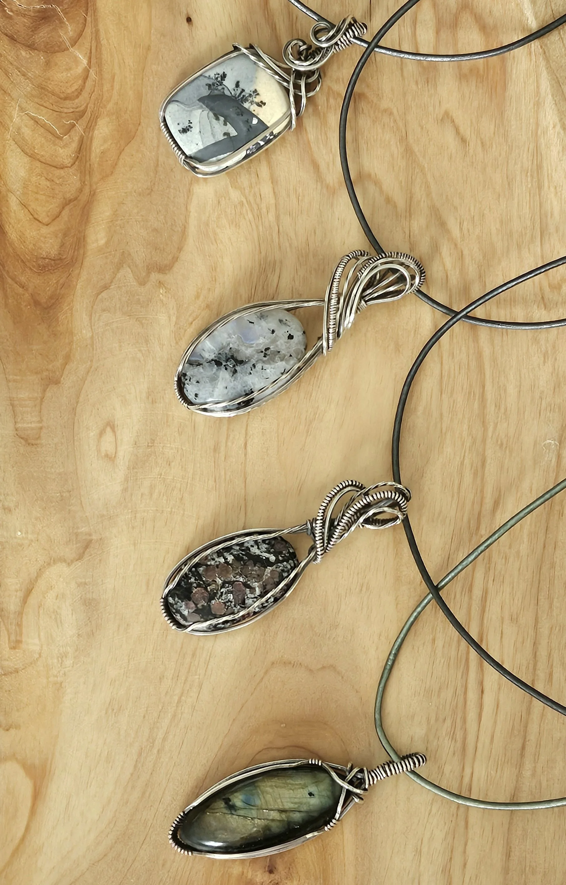 Four handmade jewelry pendants with stone or shell inlays, hanging on metal cords, displayed on a wooden surface.
