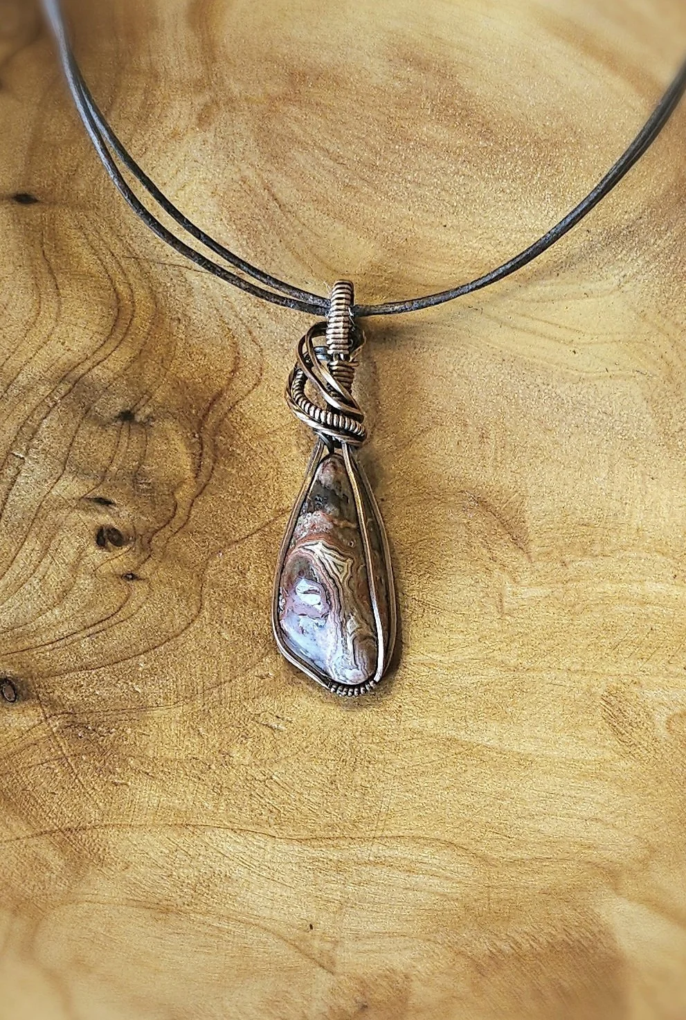 A pendant necklace with a large, teardrop-shaped stone wrapped in wire, hanging from a black cord, against a wooden background.