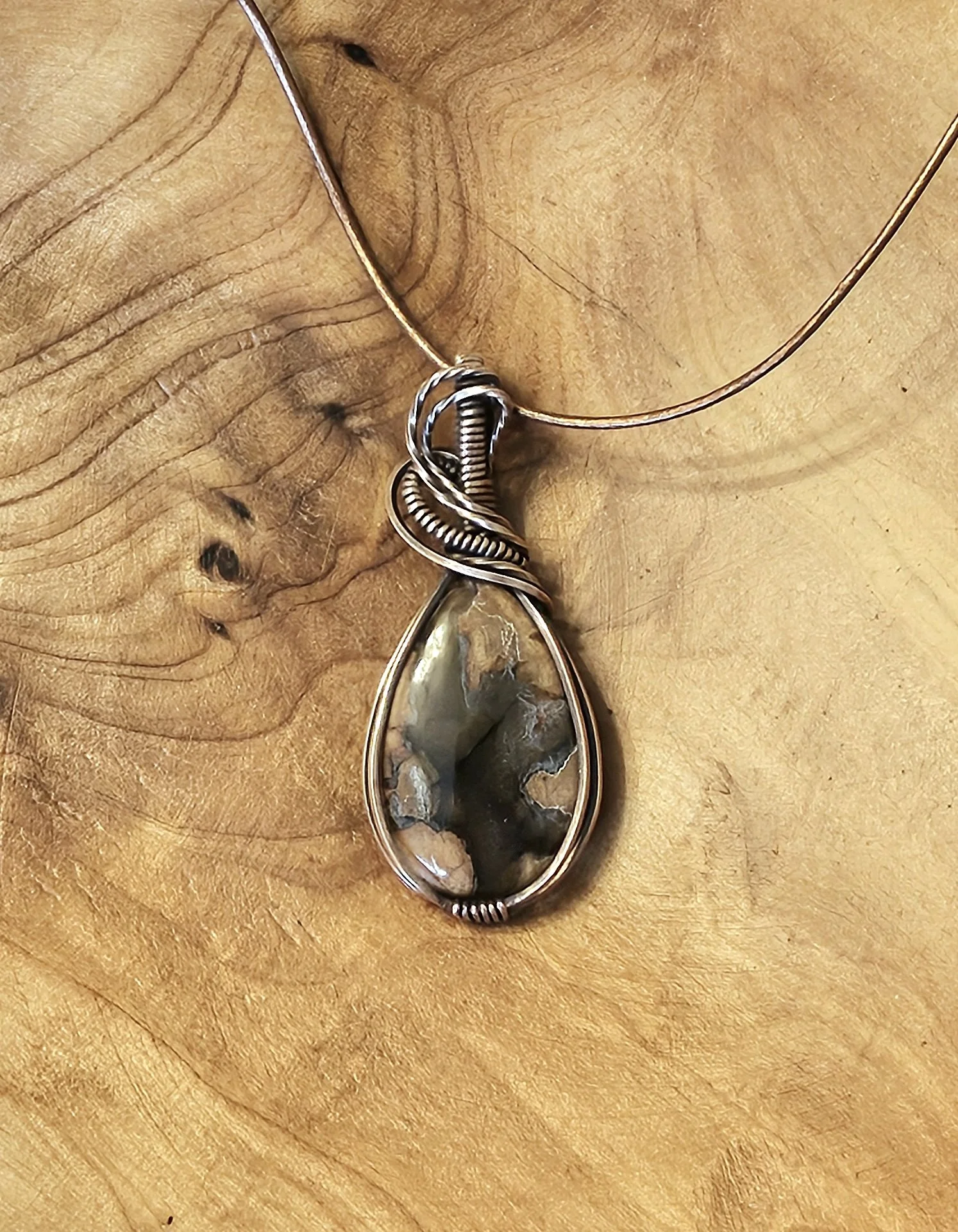 Close-up of a handmade pendant necklace with a large, teardrop-shaped stone, wrapped in wire, resting on a wooden surface.