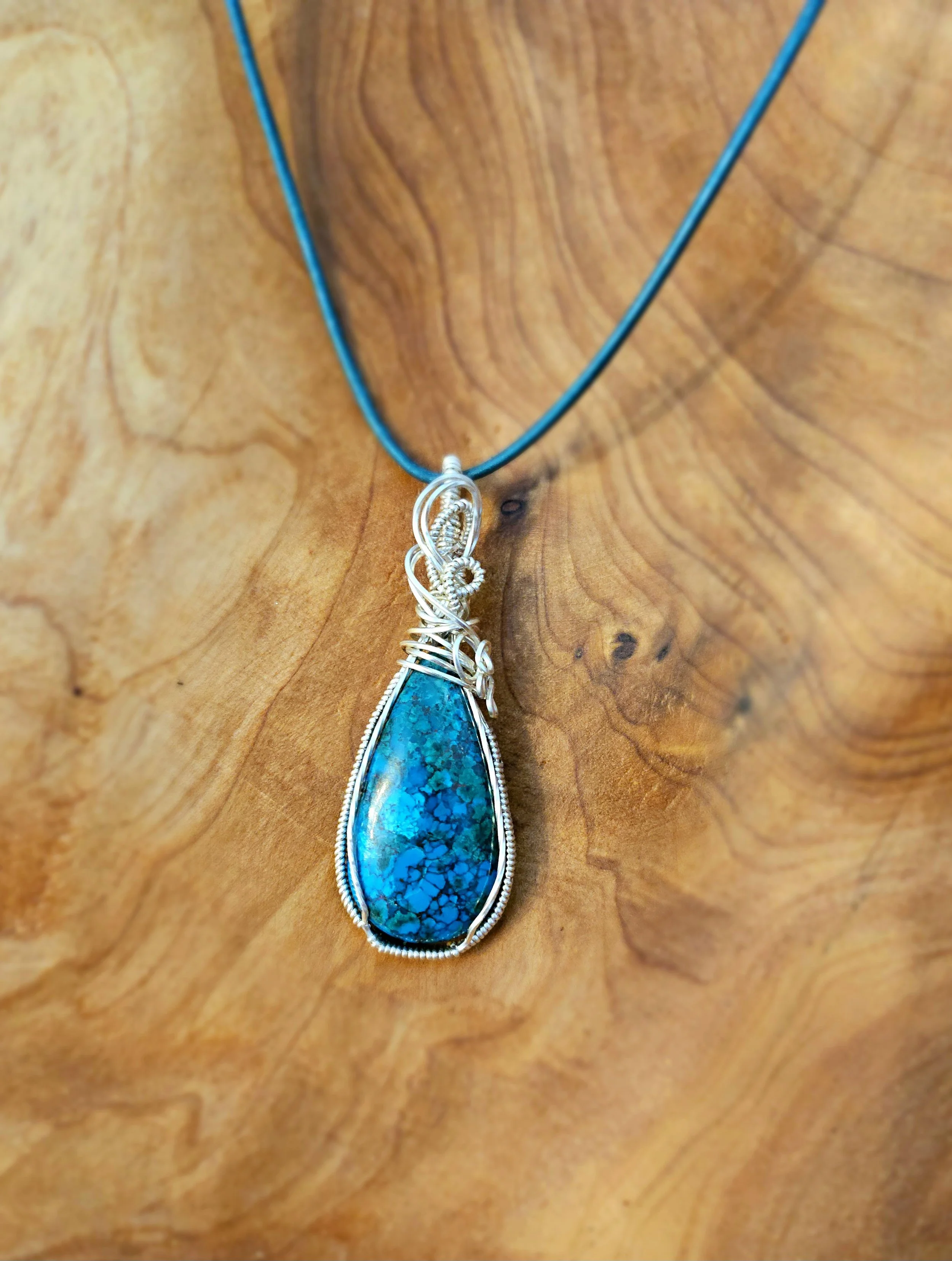 A turquoise stone pendant with silver wire wrapping on a black cord necklace displayed on a wooden surface.