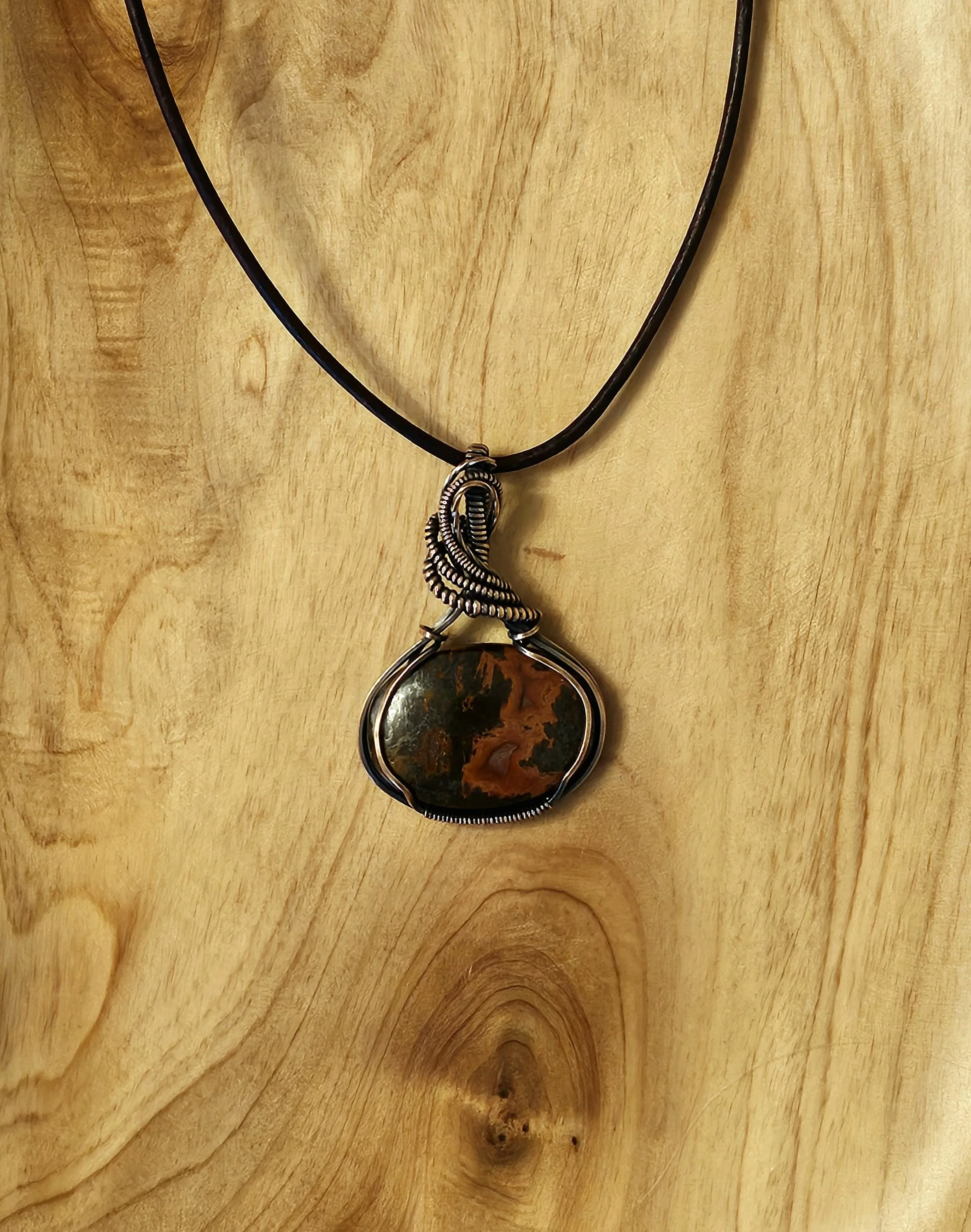 A necklace with a black cord and a large, irregularly shaped pendant featuring a multicolored stone wrapped in silver wire, hanging against a light wooden background.