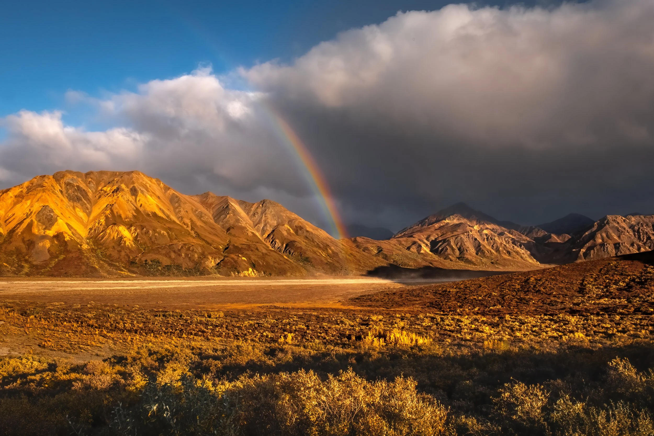 A scenic landscape of mountains with golden sunlight on their slopes, dark clouds in the sky, and a vibrant rainbow arching over the mountains, with a flat, shrub-covered plain in the foreground.