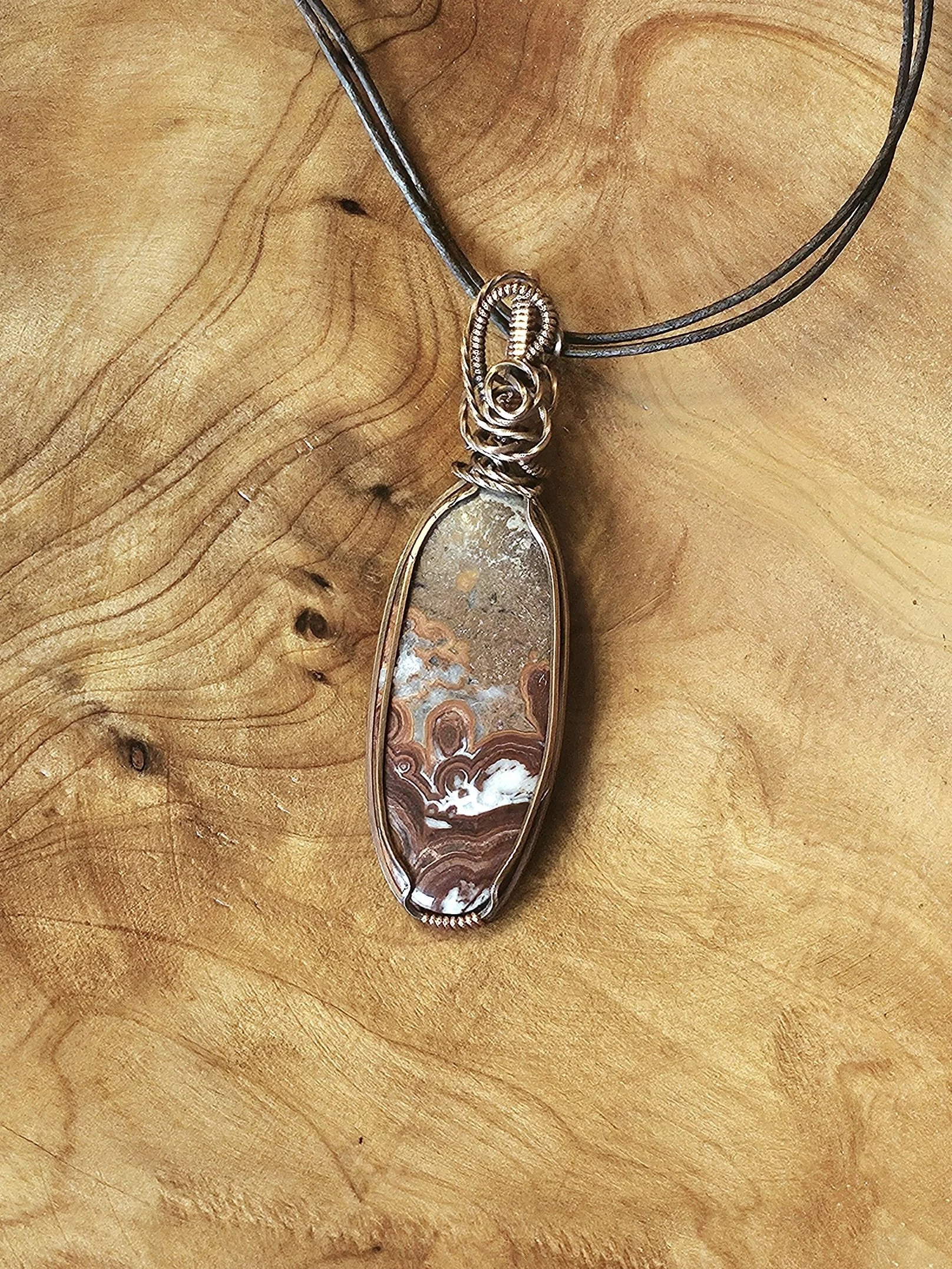 A pendant necklace featuring an oval-shaped, polished stone with brown and white banding, set in a wire-wrapped gold tone bezel, displayed on a wooden surface.