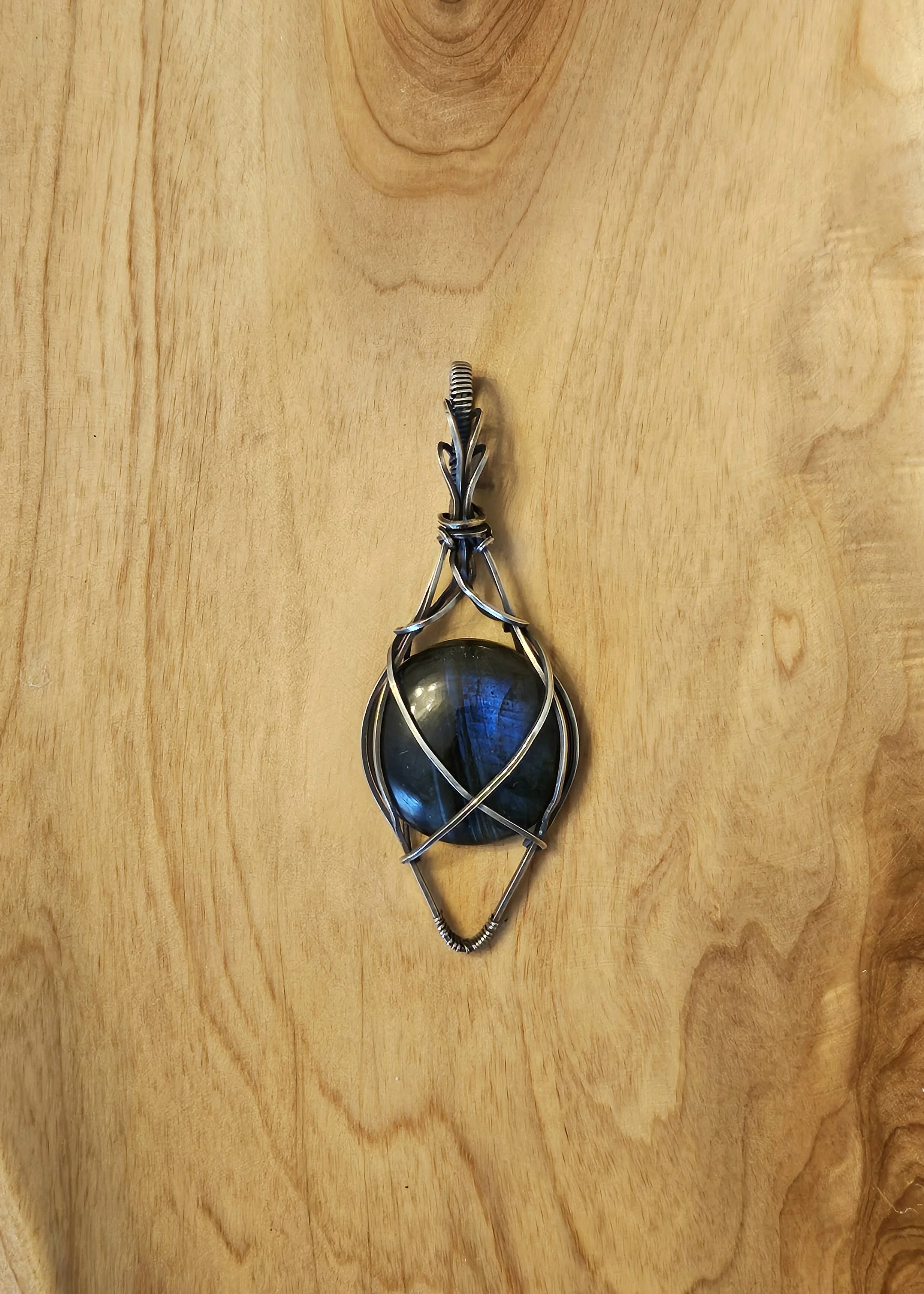 A pendant with a dark, reflective gemstone wrapped in silver wire, hanging against a light wooden background.