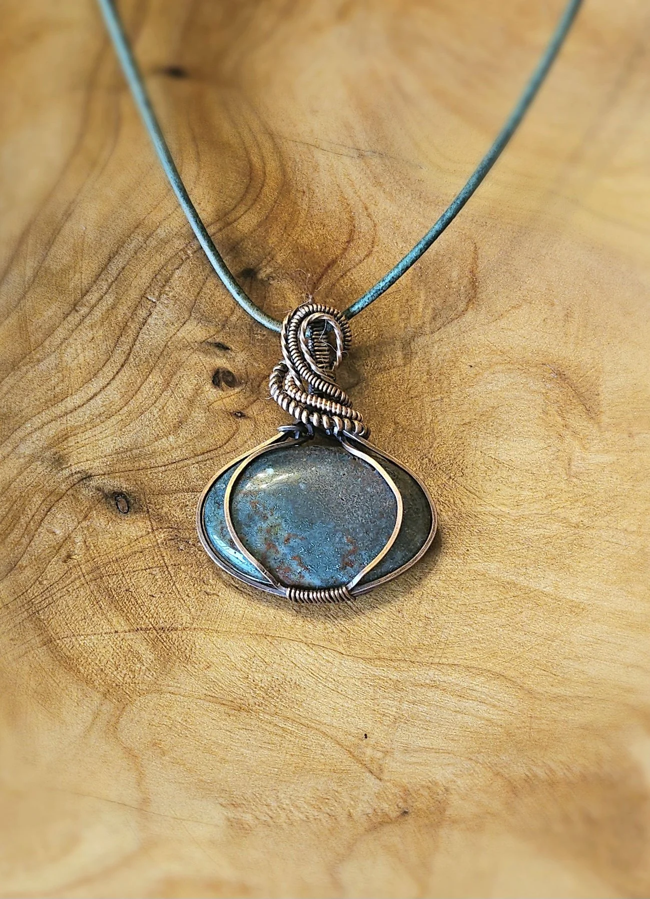 A silver necklace with a pendant featuring a round, bluish-green stone, hanging on a black cord against a wooden background.