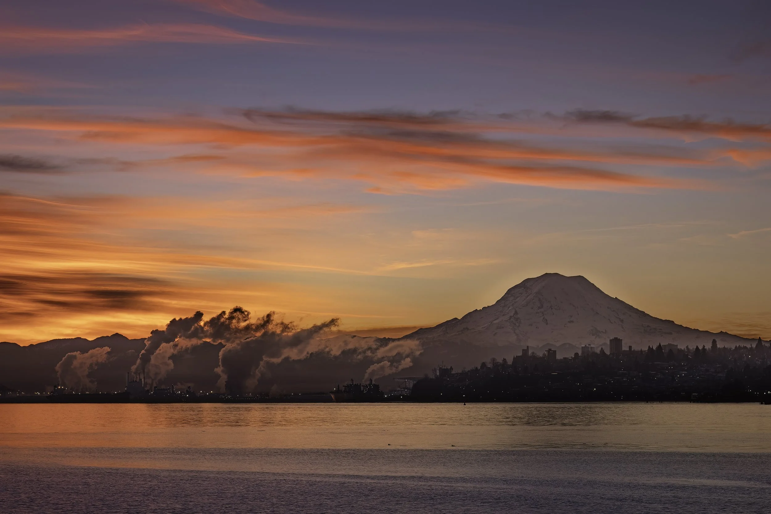 Sunset over a mountain with a snow-capped peak, possibly Mount Fuji, with an industrial area and smokestacks emitting smoke in the foreground, and water in the front.