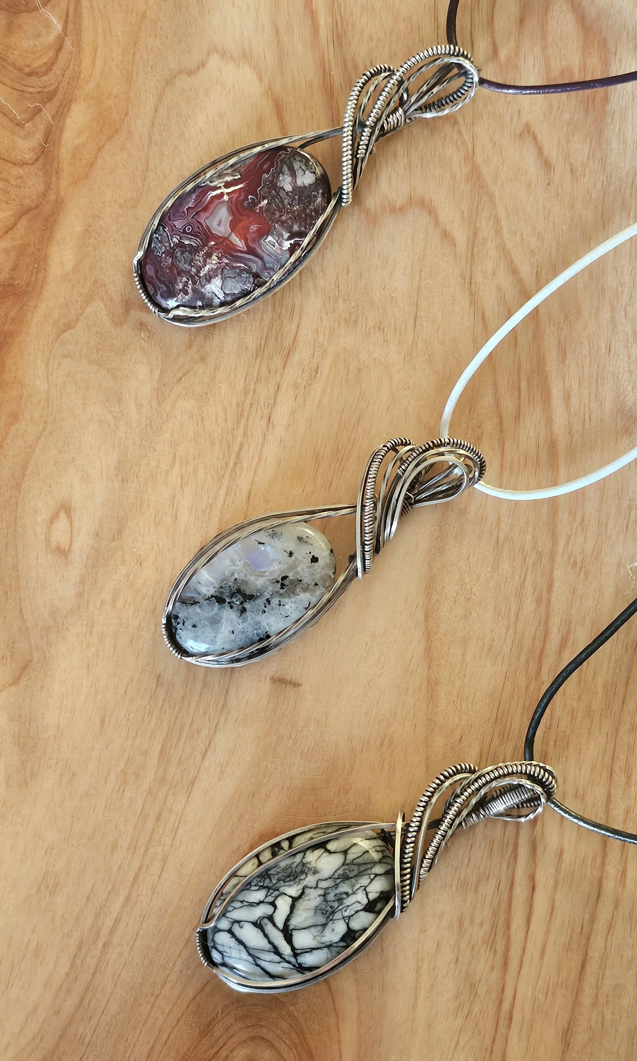 Three wire-wrapped gemstone necklaces with oval stones in red, gray, and black and white patterns, laid out on a wooden surface.
