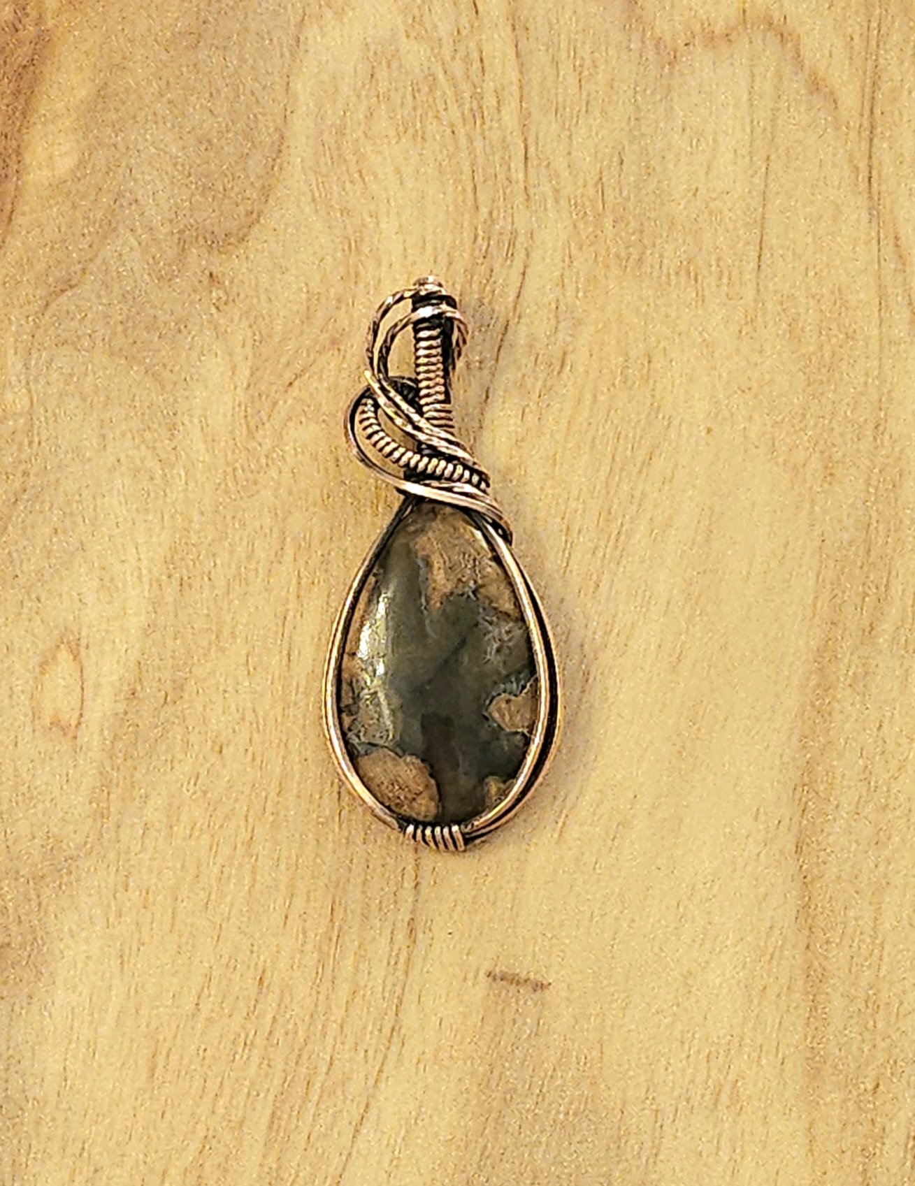 A teardrop-shaped pendant with a dark, marbled stone, wrapped in gold-colored wire, on a wooden surface.