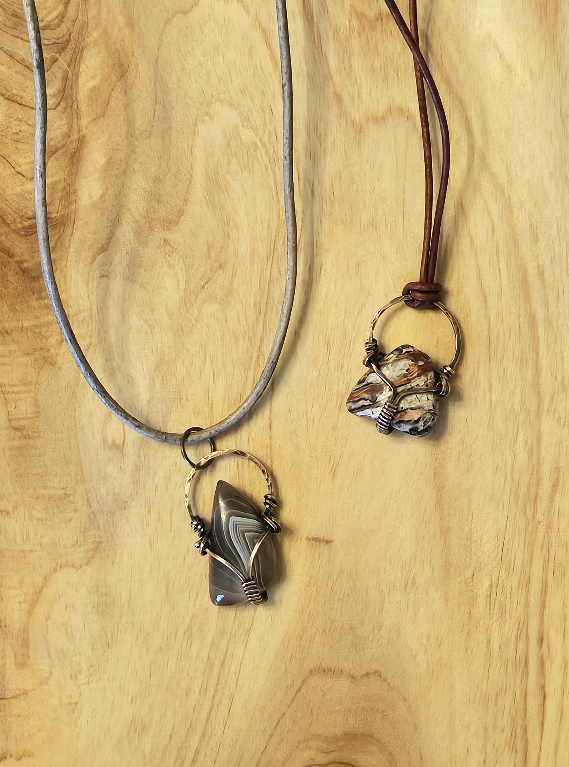 Two necklaces with handmade stone pendants on leather cords, hanging on a wooden surface.