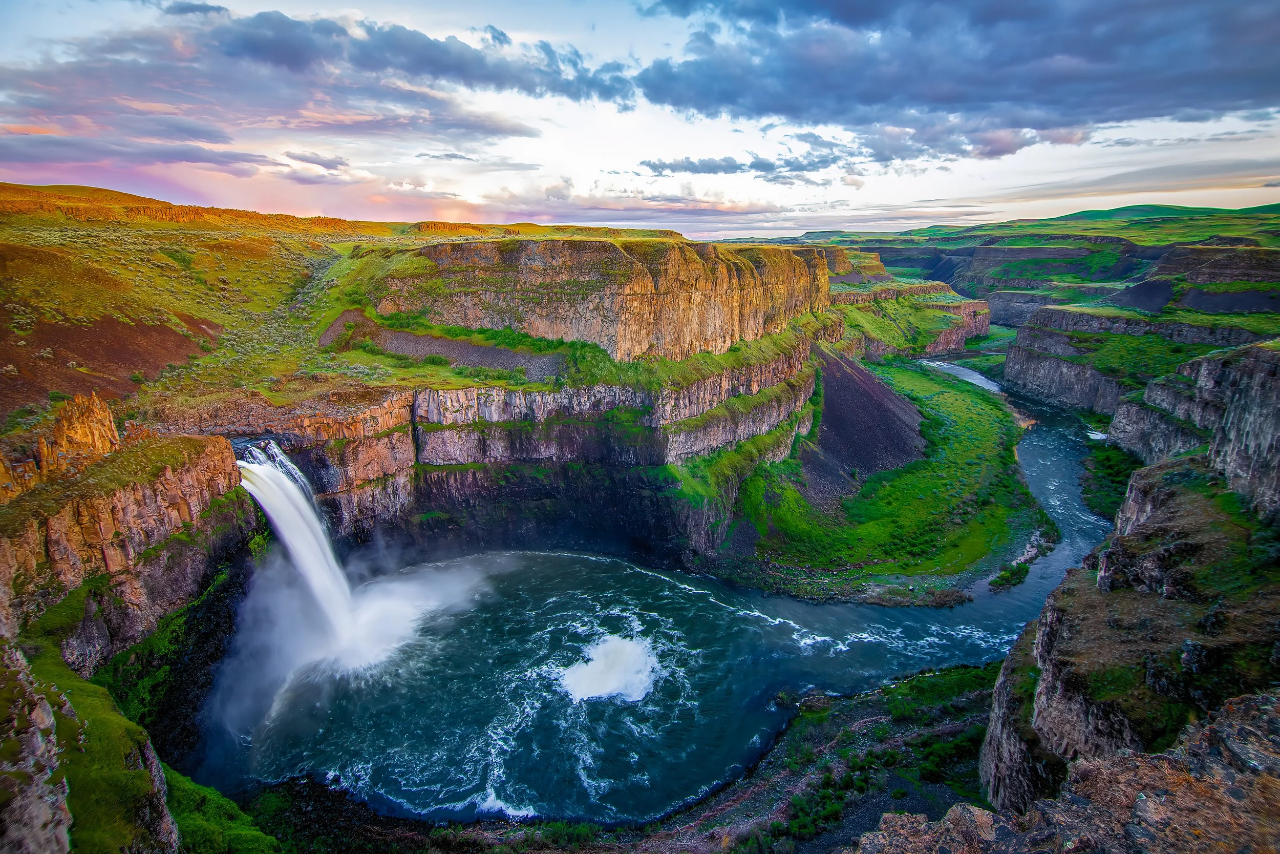 Scenic view of a waterfall in a canyon with green cliffs, a river, and a colorful sky with clouds during sunset.
