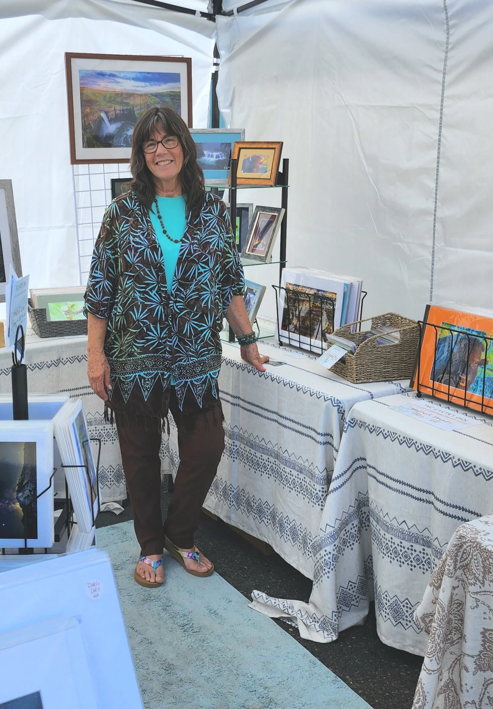 A woman standing in front of an art display booth, smiling, wearing glasses, a tropical patterned top and dark pants, with artwork and photos on tables and shelves behind her.