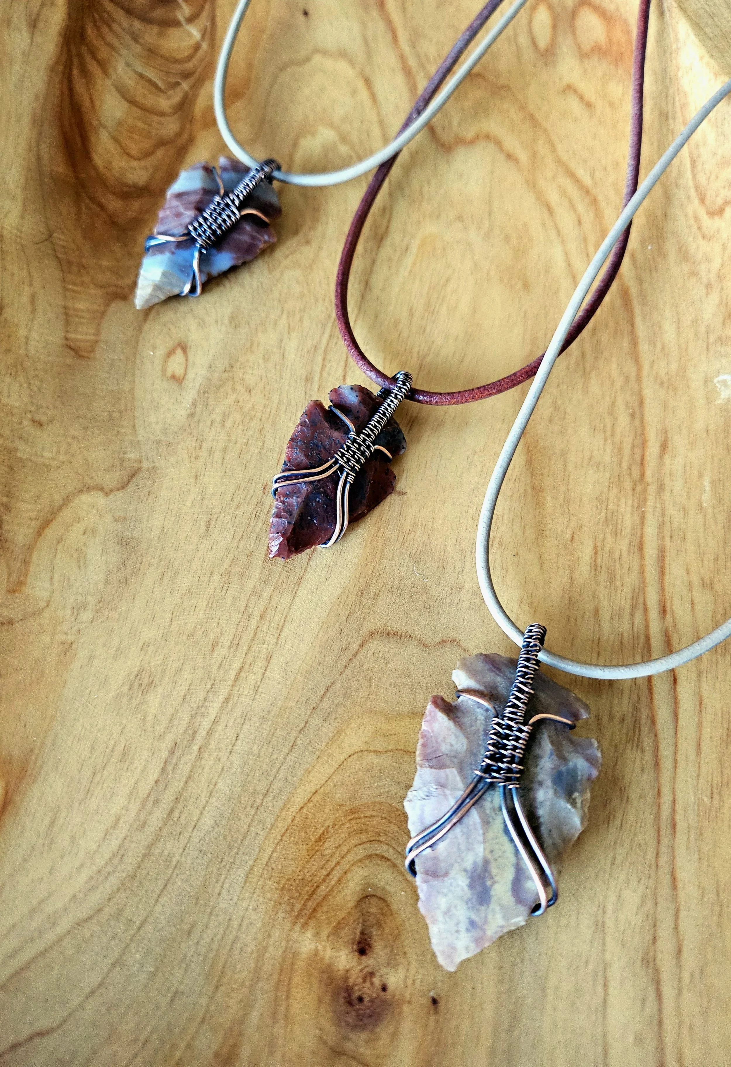 Three necklaces with large arrowhead pendants wrapped in wire, displayed on a wooden surface.