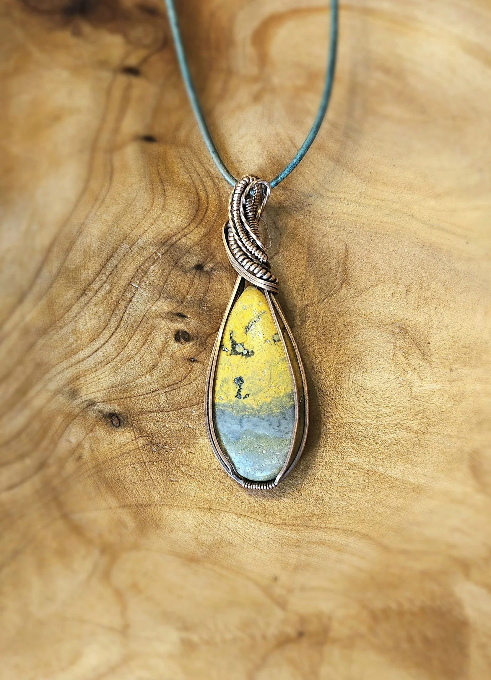 A pendant necklace with a yellow, gray, and blue stone wrapped in silver wire, hanging from a light blue cord on a wooden surface.