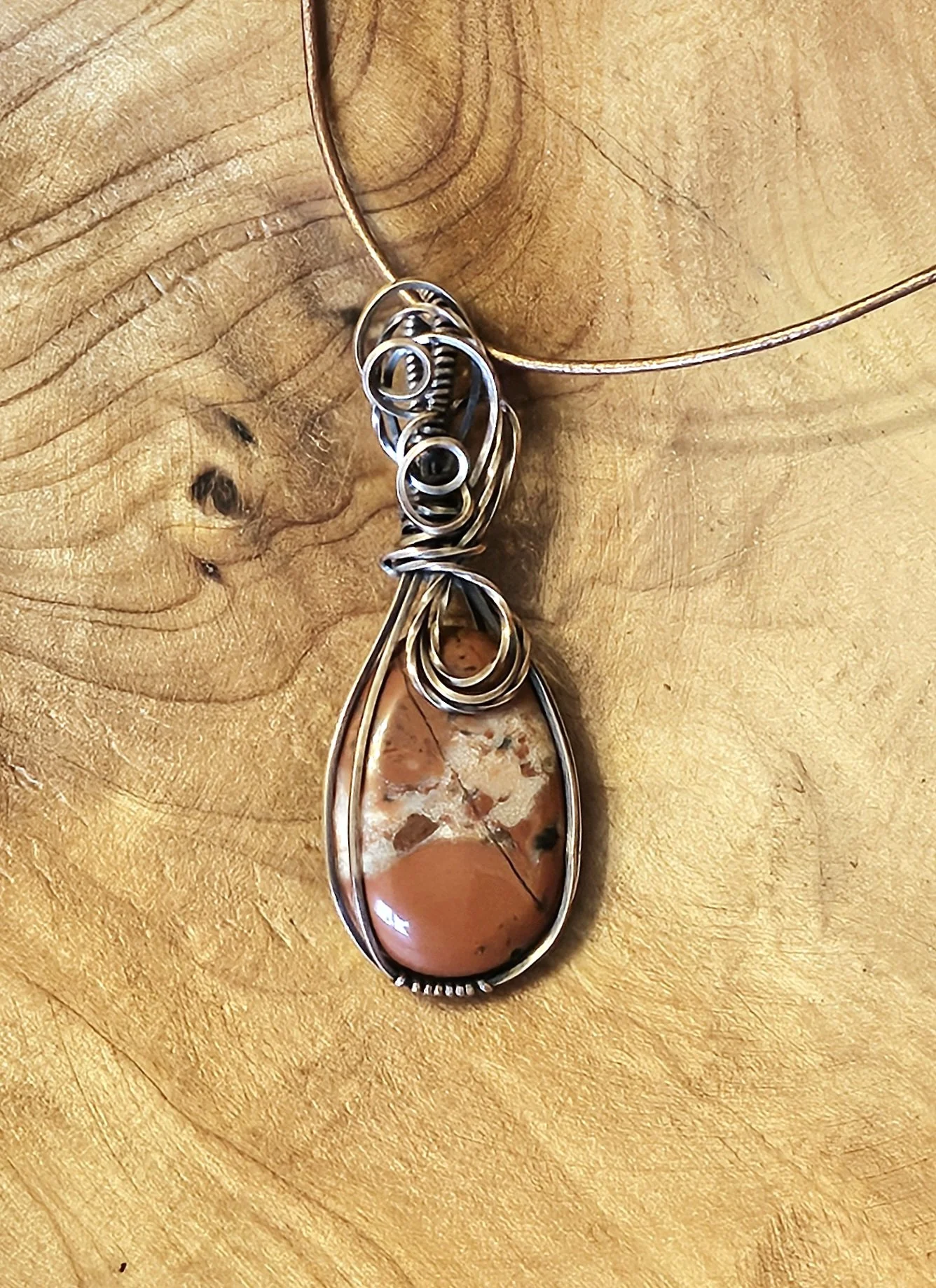A piece of jewelry with a large teardrop-shaped stone encased in wire, hanging from a wire necklace, placed on a wooden surface.