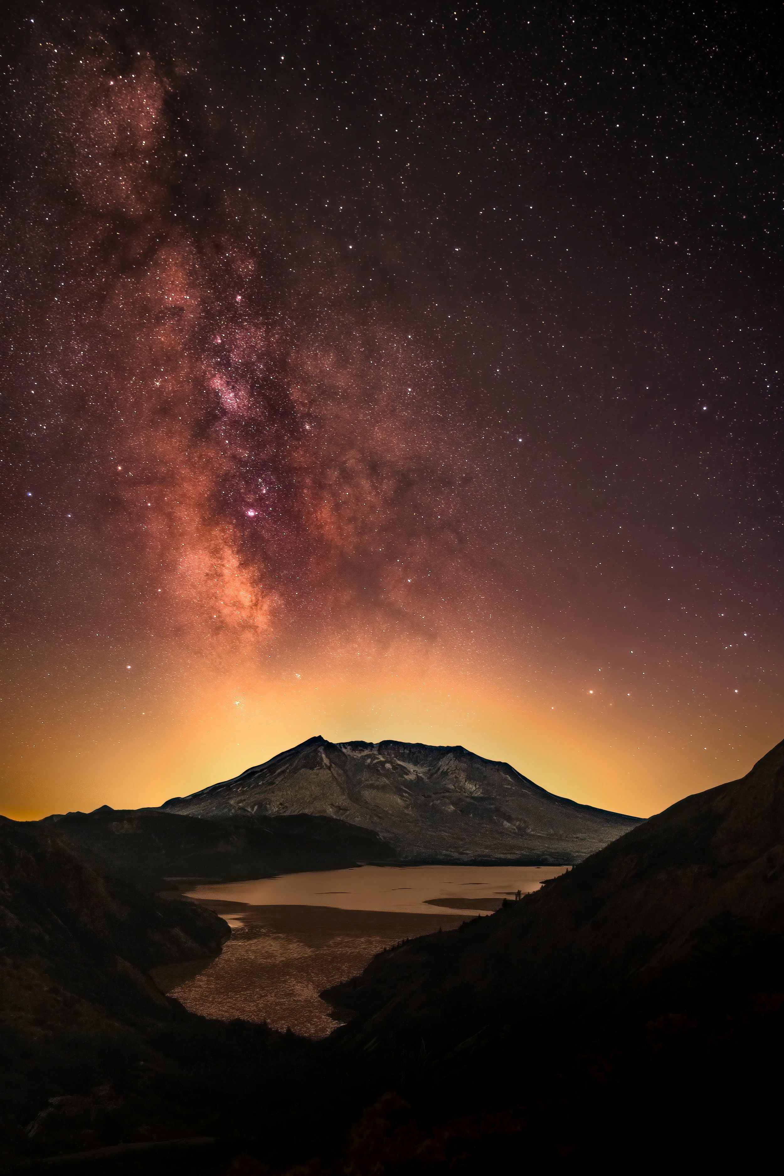 Night sky filled with stars and the Milky Way galaxy over a mountain and lake at dusk or dawn.