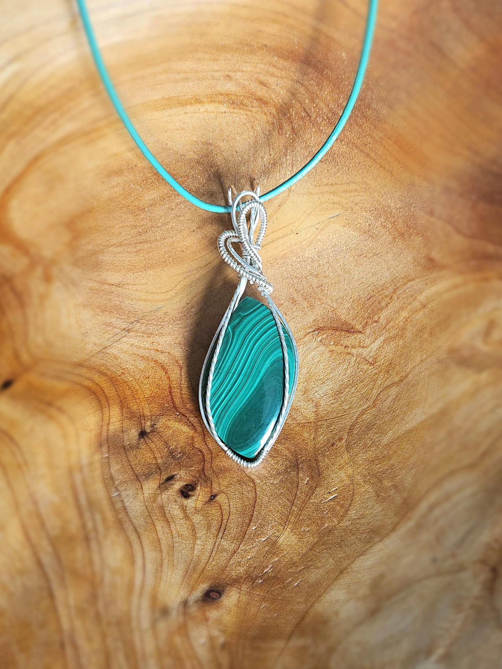 A turquoise gemstone pendant with silver wire wrapping, hanging on a turquoise cord, set against a wooden background.