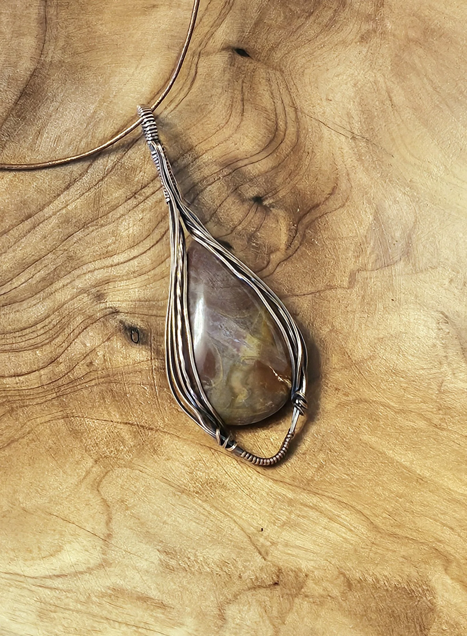 A teardrop-shaped pendant with a multicolored stone in a wire-wrapped silver setting, hanging on a matching silver chain against a textured wooden background.
