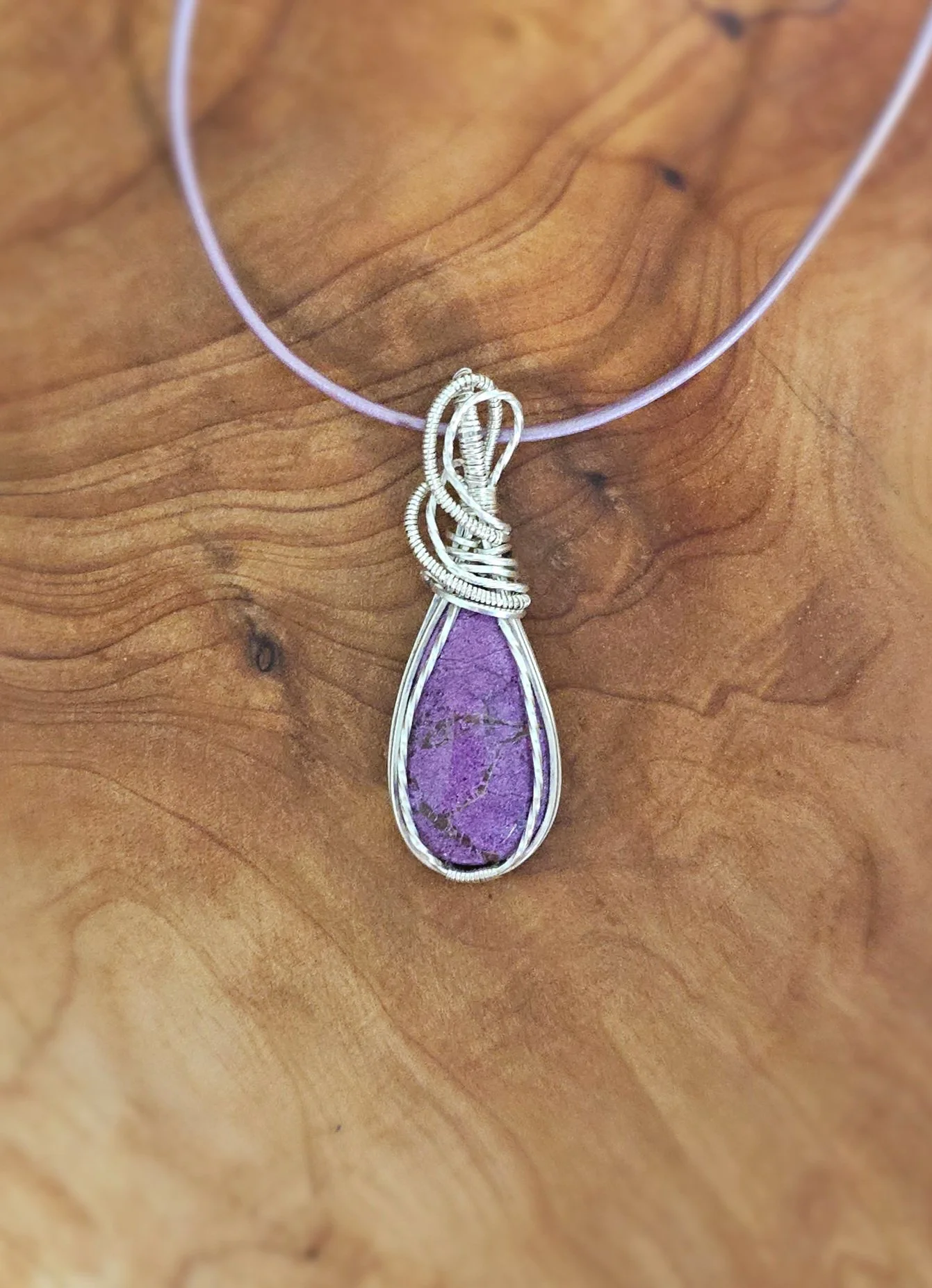A silver wire-wrapped necklace with a purple teardrop-shaped gemstone pendant resting on a wooden surface.