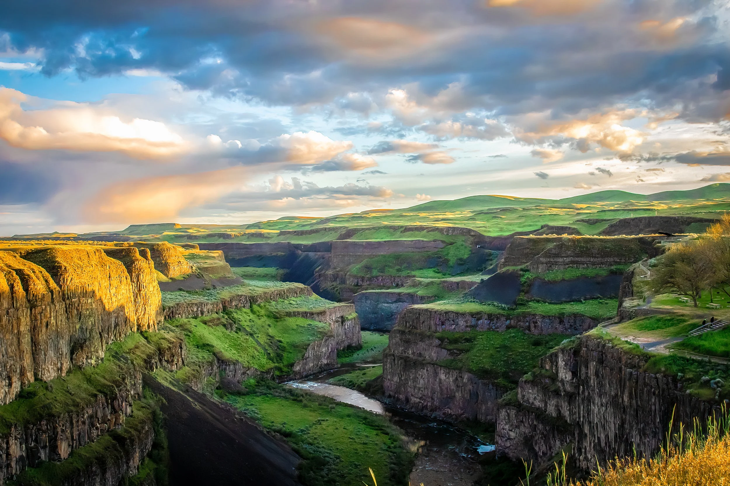 Sunset over a deep canyon with green grass and rocky cliffs, river flowing at the bottom, cloudy sky.