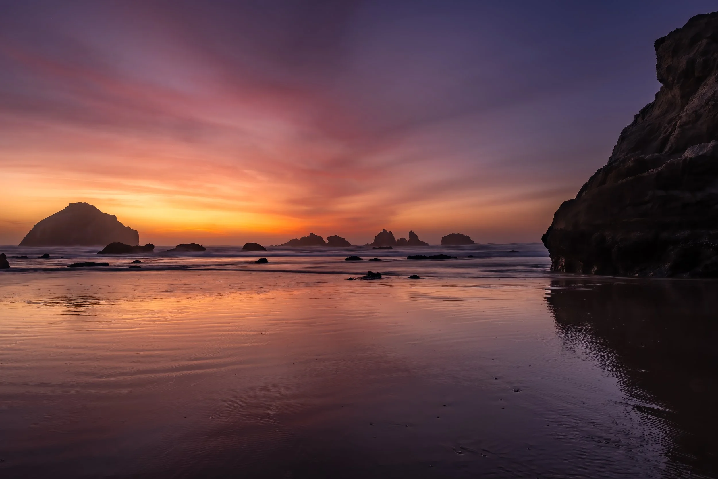 Sunset over a rocky beach with calm water reflecting the colorful sky and silhouetted rocks and cliffs.