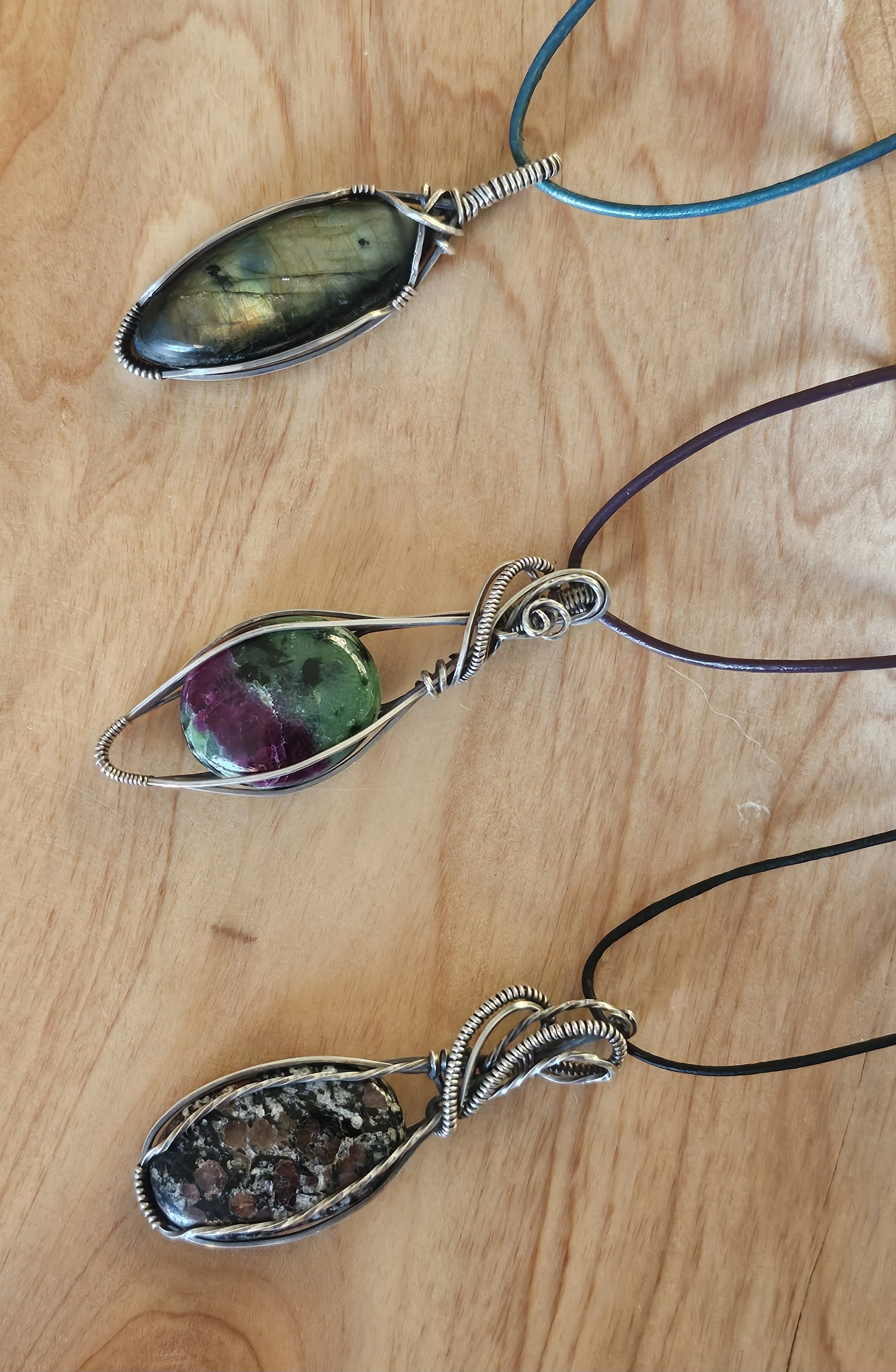 Three wire-wrapped pendants with large stones, each attached to a cord, arranged on a wooden surface.