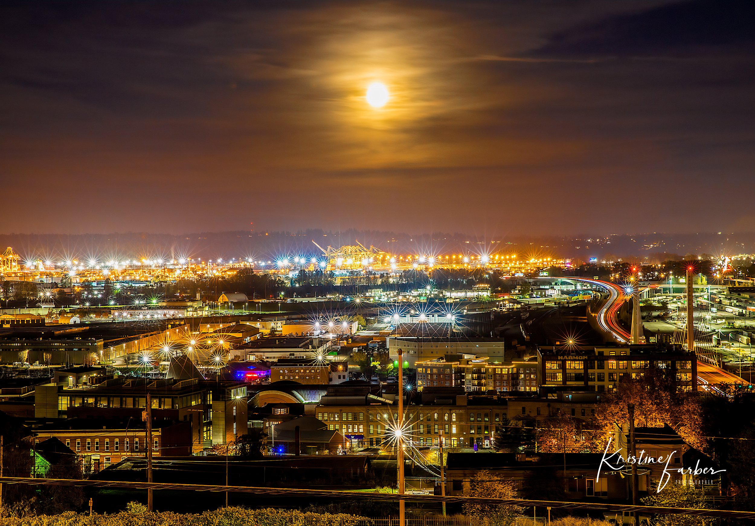 Nighttime cityscape with illuminated buildings, bright streetlights, and a full moon in the sky over a city with industrial and residential areas.