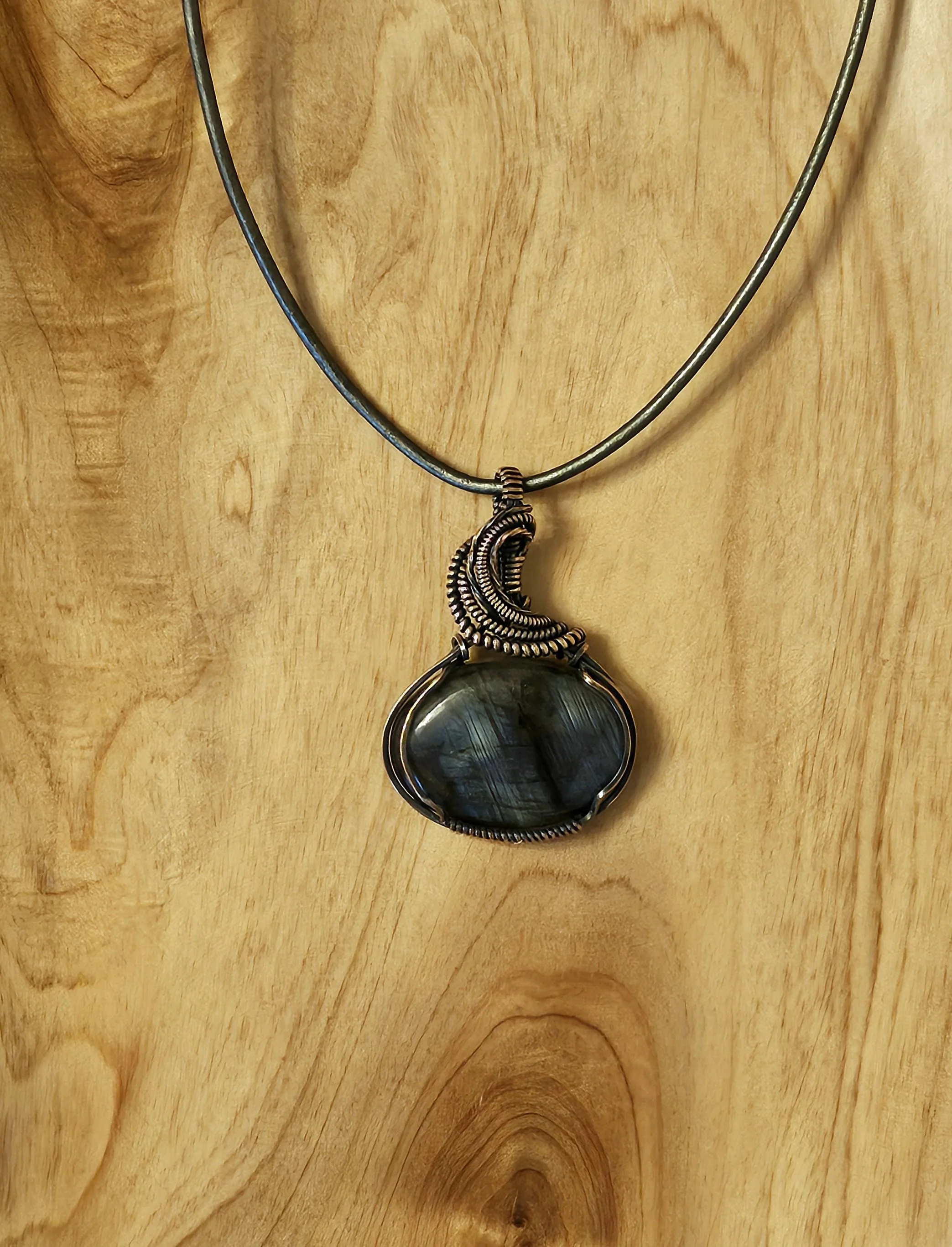 A necklace with a large, polished black stone pendant hanging from a metallic, intricately designed crescent-shaped bail on a thin, metallic cord against a wooden background.