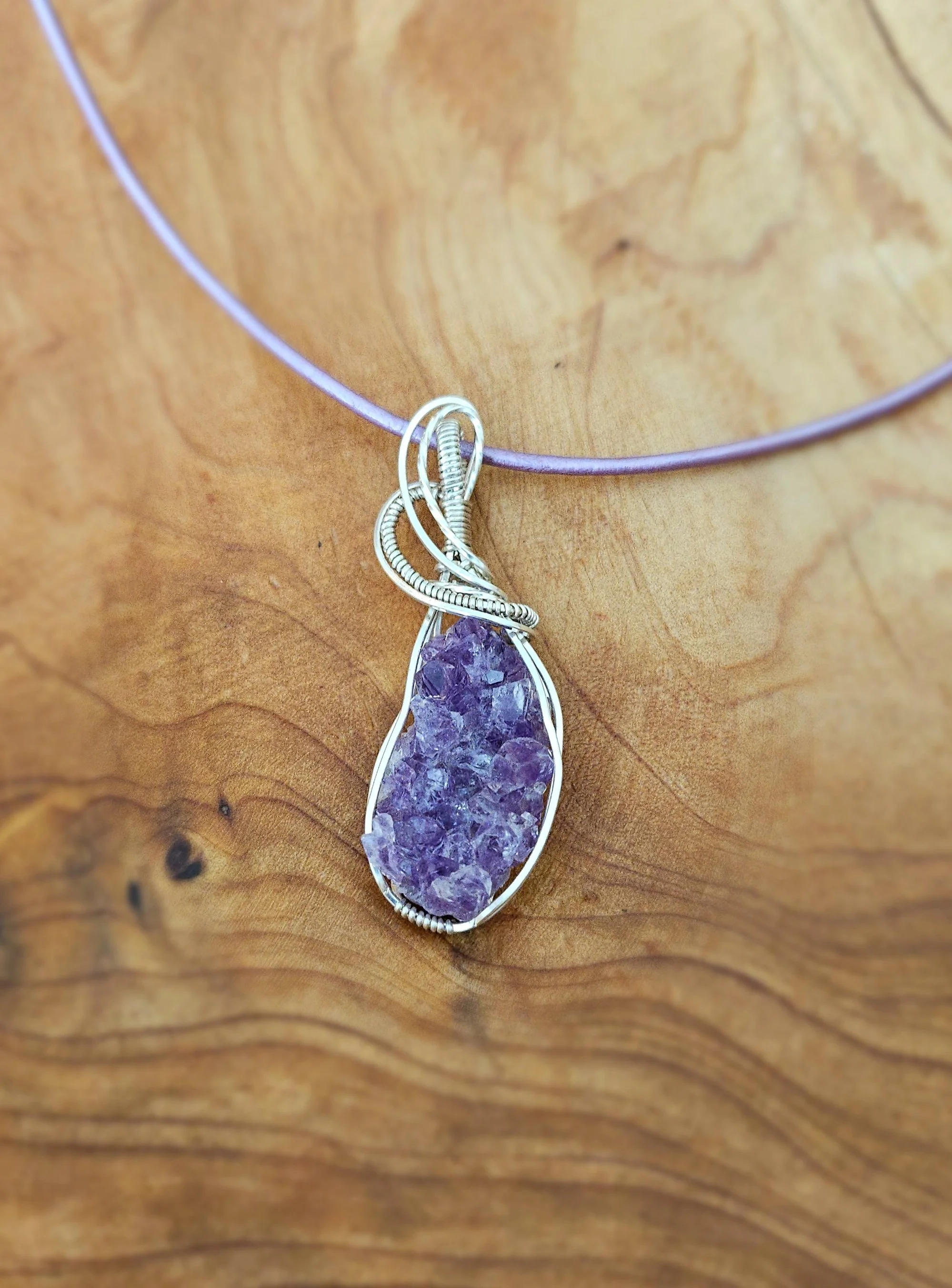 A jewelry pendant necklace with a large, irregularly shaped purple crystal set in a silver wire cage, hanging from a thin purple cord on a wooden surface.