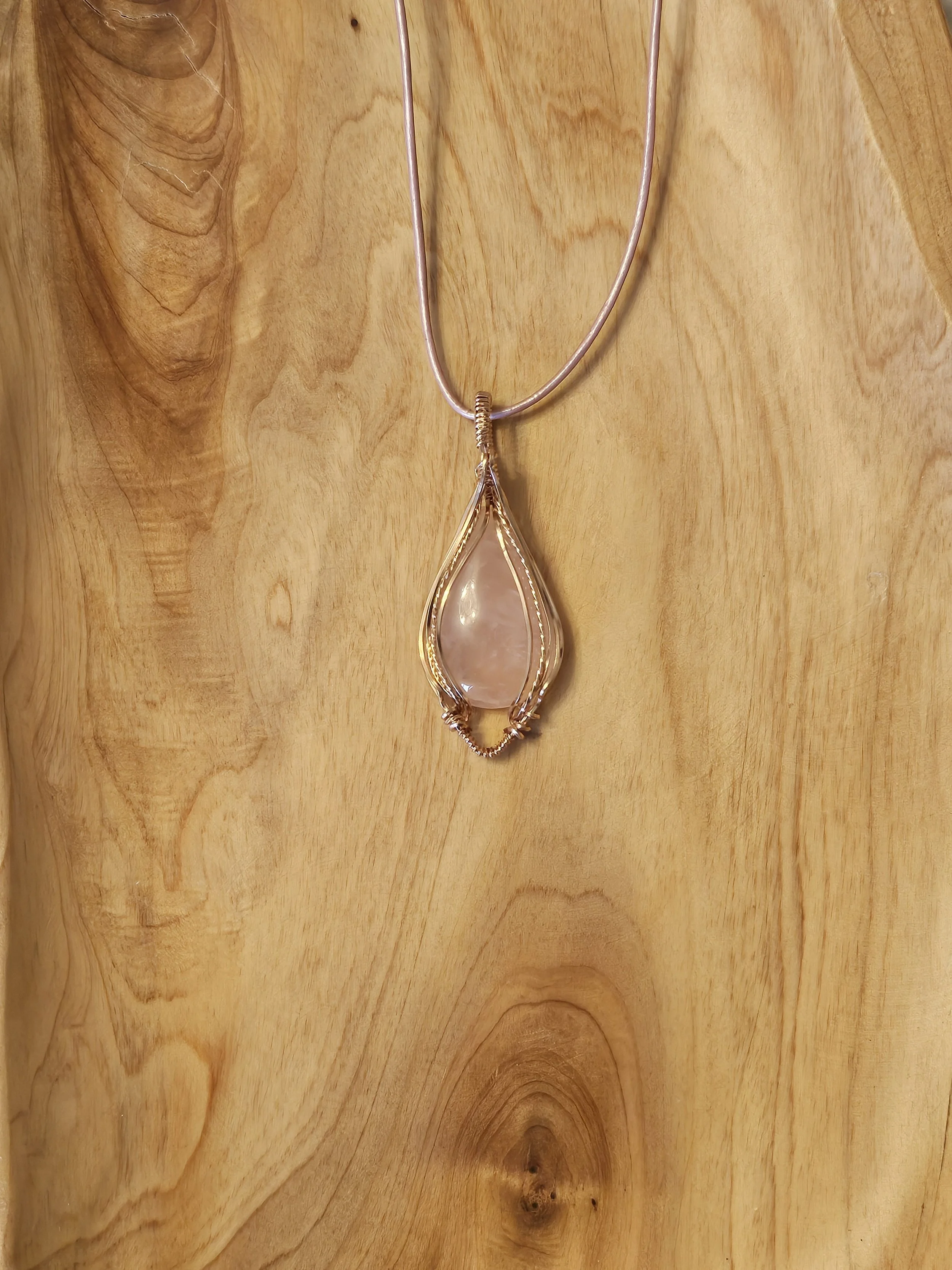 A pendant necklace with a large teardrop-shaped stone encased in a gold wire setting, hanging from a thin silver chain, displayed against a wooden background.