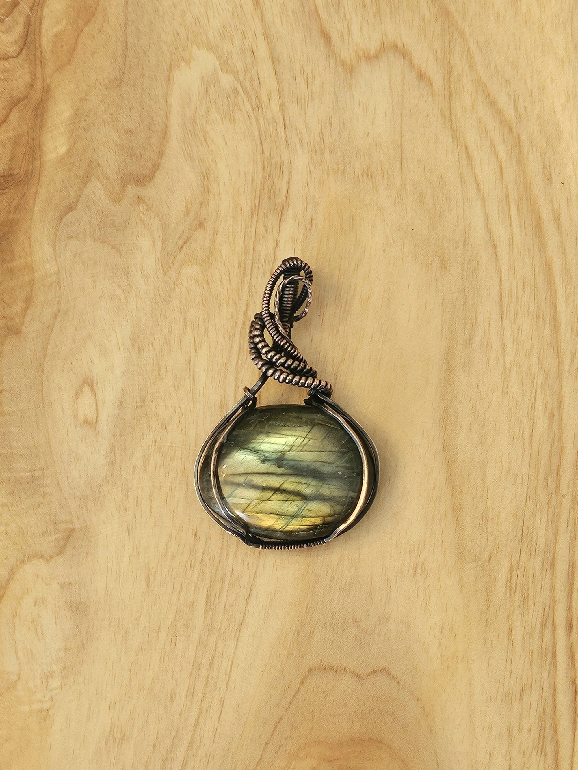 A pendant with a large, oval-shaped, iridescent stone wrapped in wire, resting on a light wooden surface.