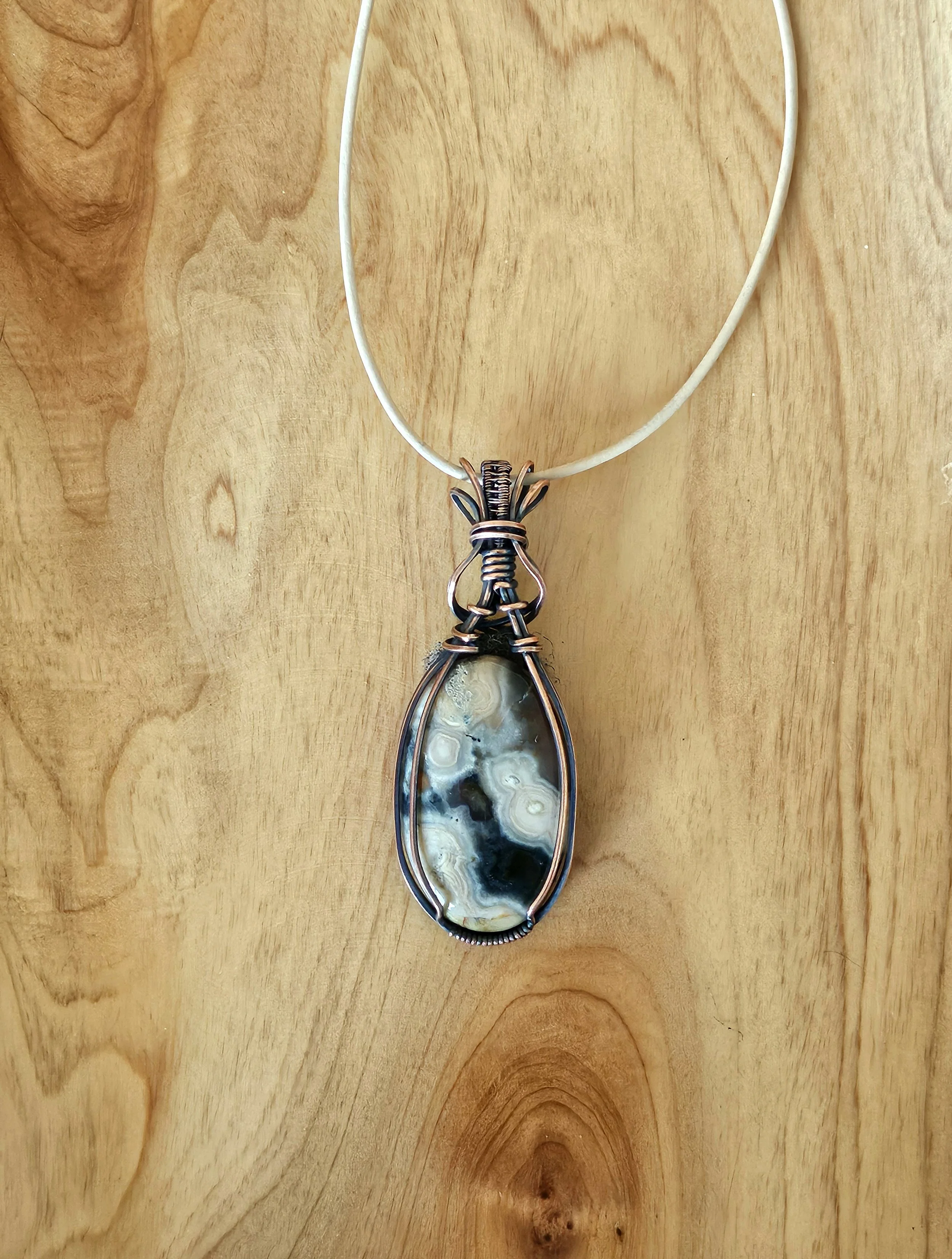 Handcrafted wire-wrapped pendant with a large, polished, multi-colored stone on a white cord necklace, displayed on a light wooden surface.