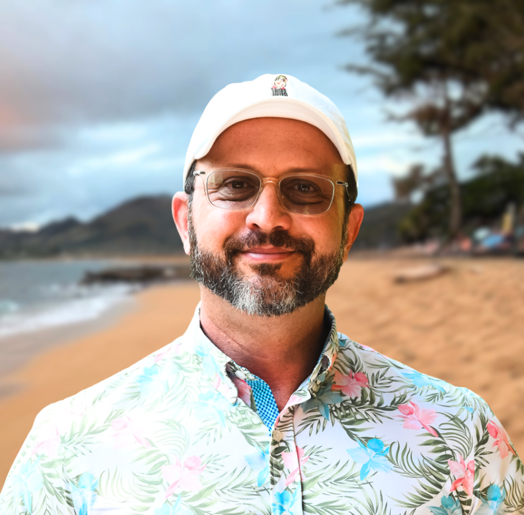 A smiling, bald man with a goatee, wearing a bright pink shirt, standing outdoors with a body of water and a blue sky in the background.