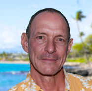 Close-up of a man outdoors with palm trees and a pool in the background.