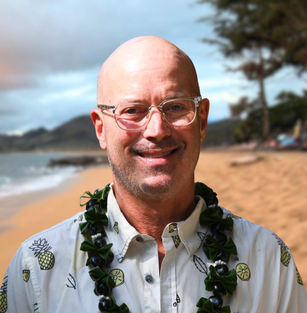 Smiling man with glasses and a patterned shirt outdoors near water and trees