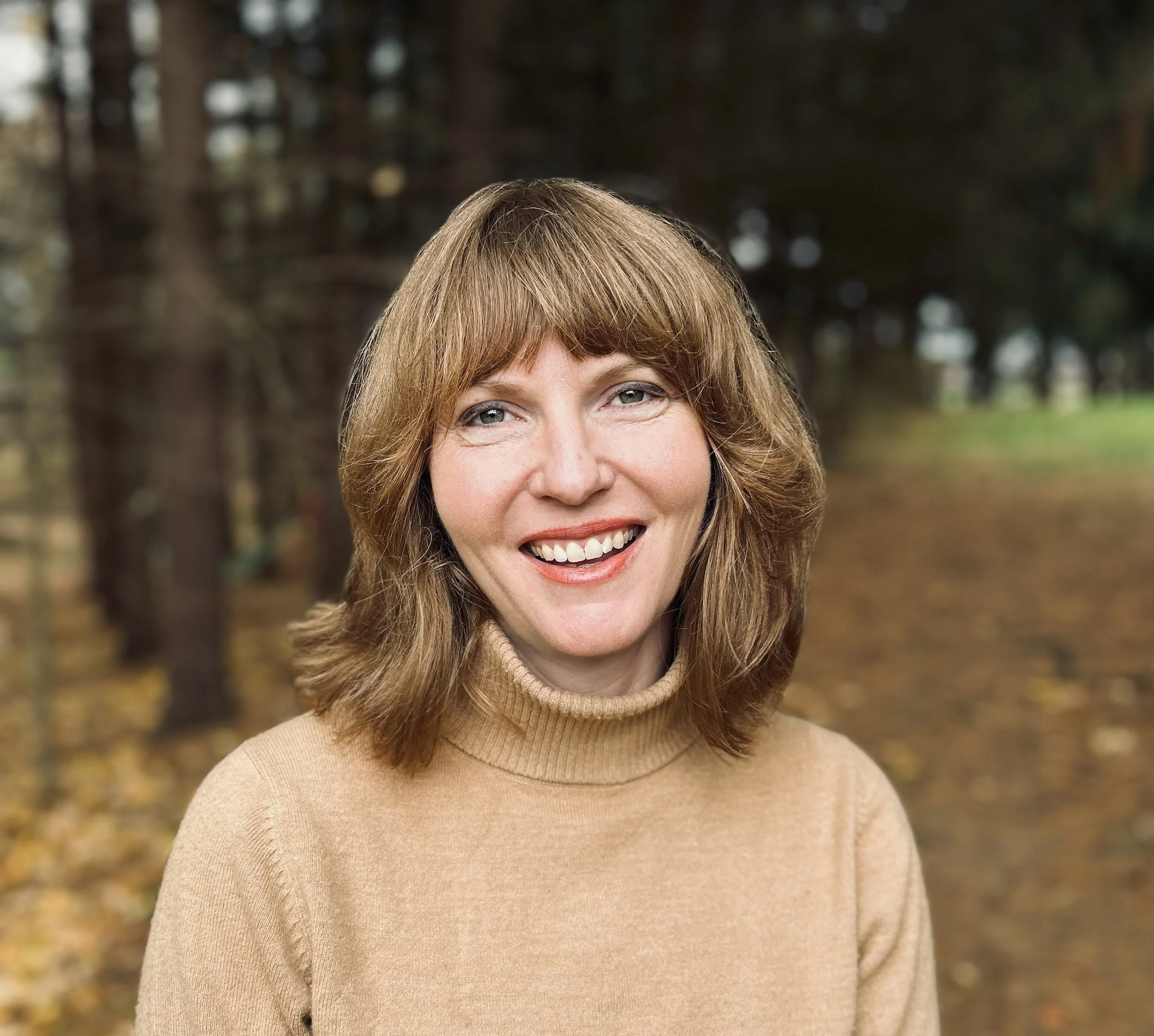 Therapist, Diana Dunn with shoulder-length light brown hair smiling, wearing a beige turtleneck sweater, standing outdoors in a forest with trees and fallen leaves.