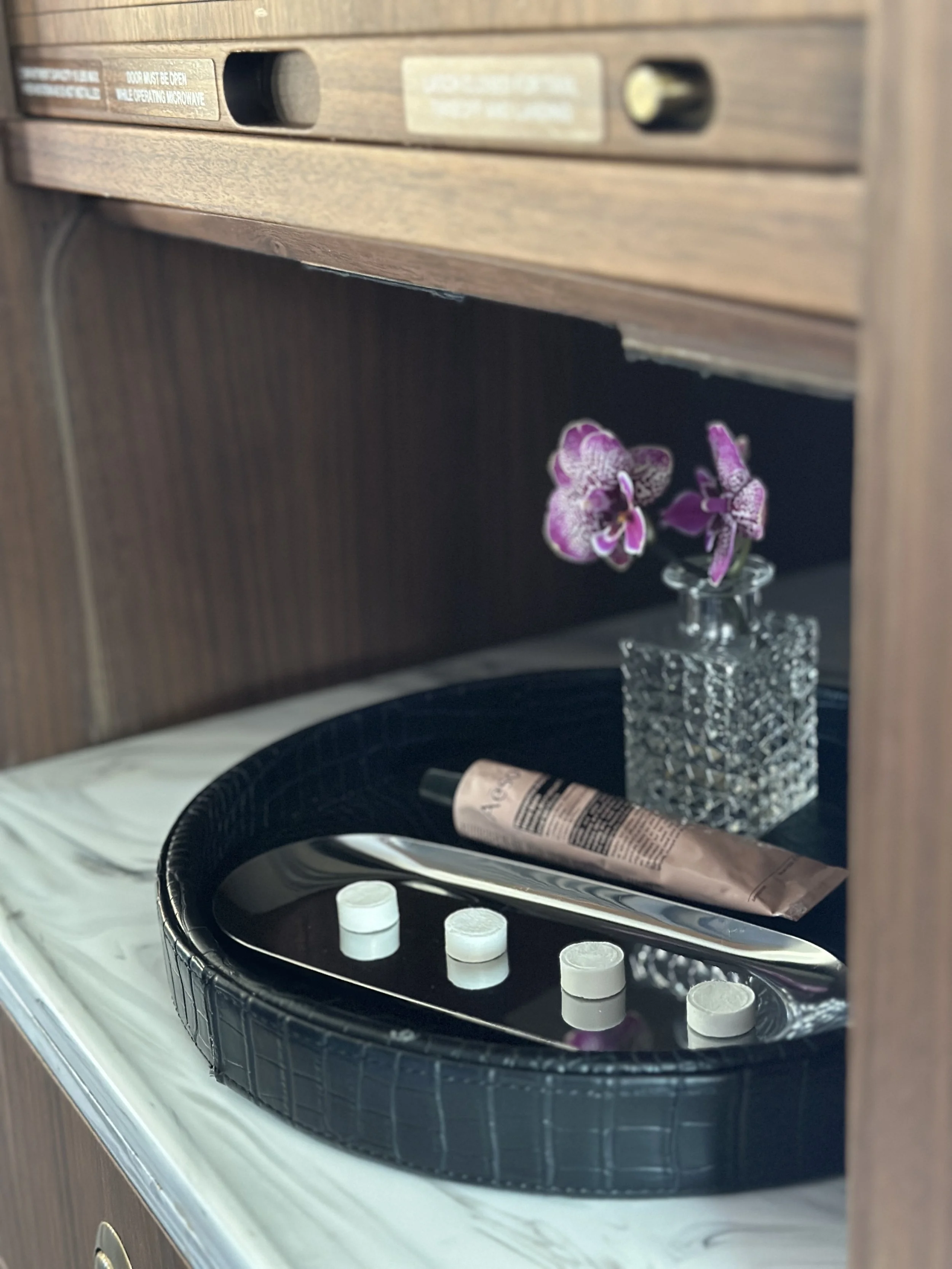 A black tray with four white tablets, a tube, and a metal tongue cleaner on a white marble surface. Behind the tray is a small glass vase with pink and purple orchids inside.
