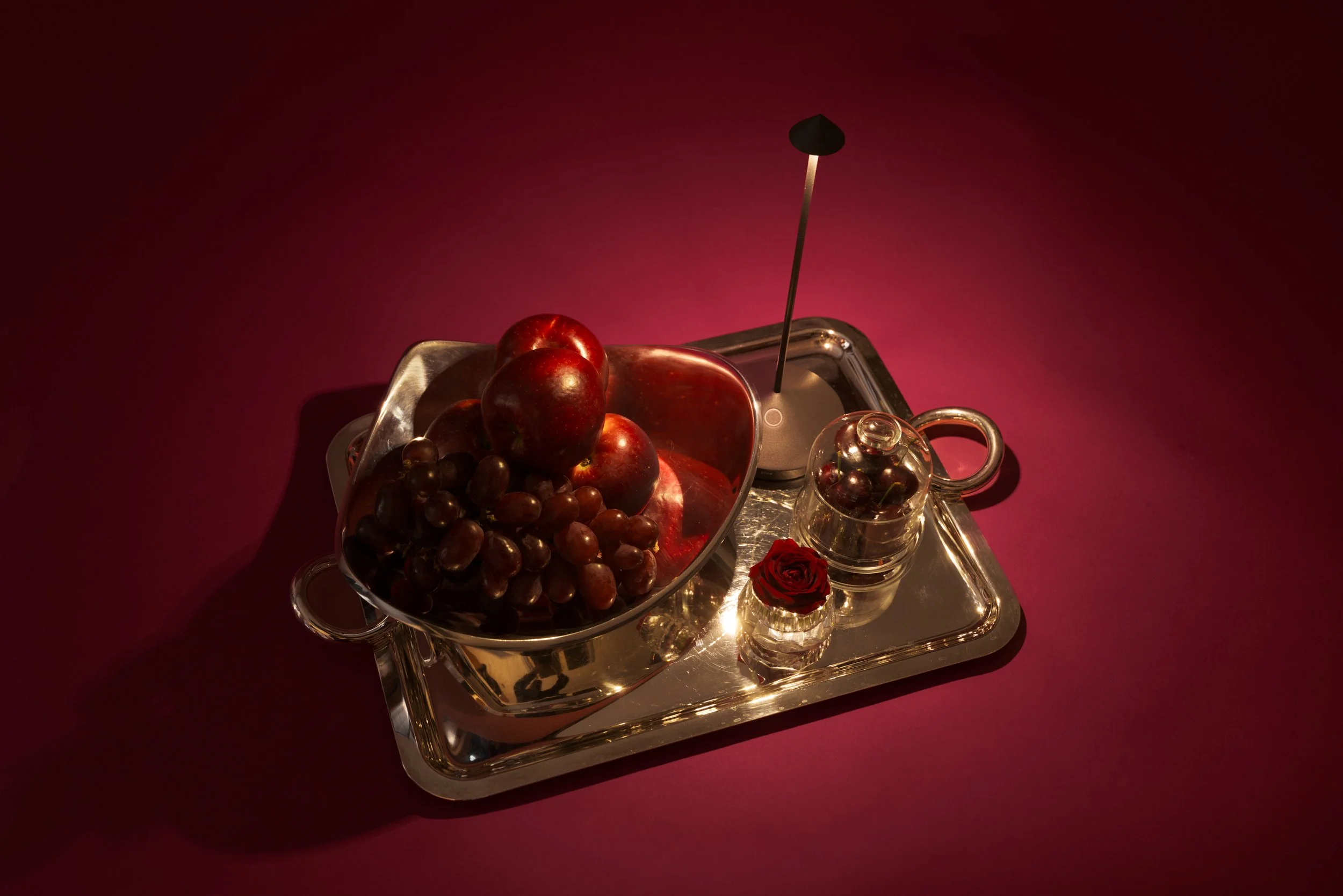 Silver tray with a silver bowl of apples and grapes, a small glass pitcher with grapes, a small glass of water with a red rose, and an oil lamp, all set against a red background.