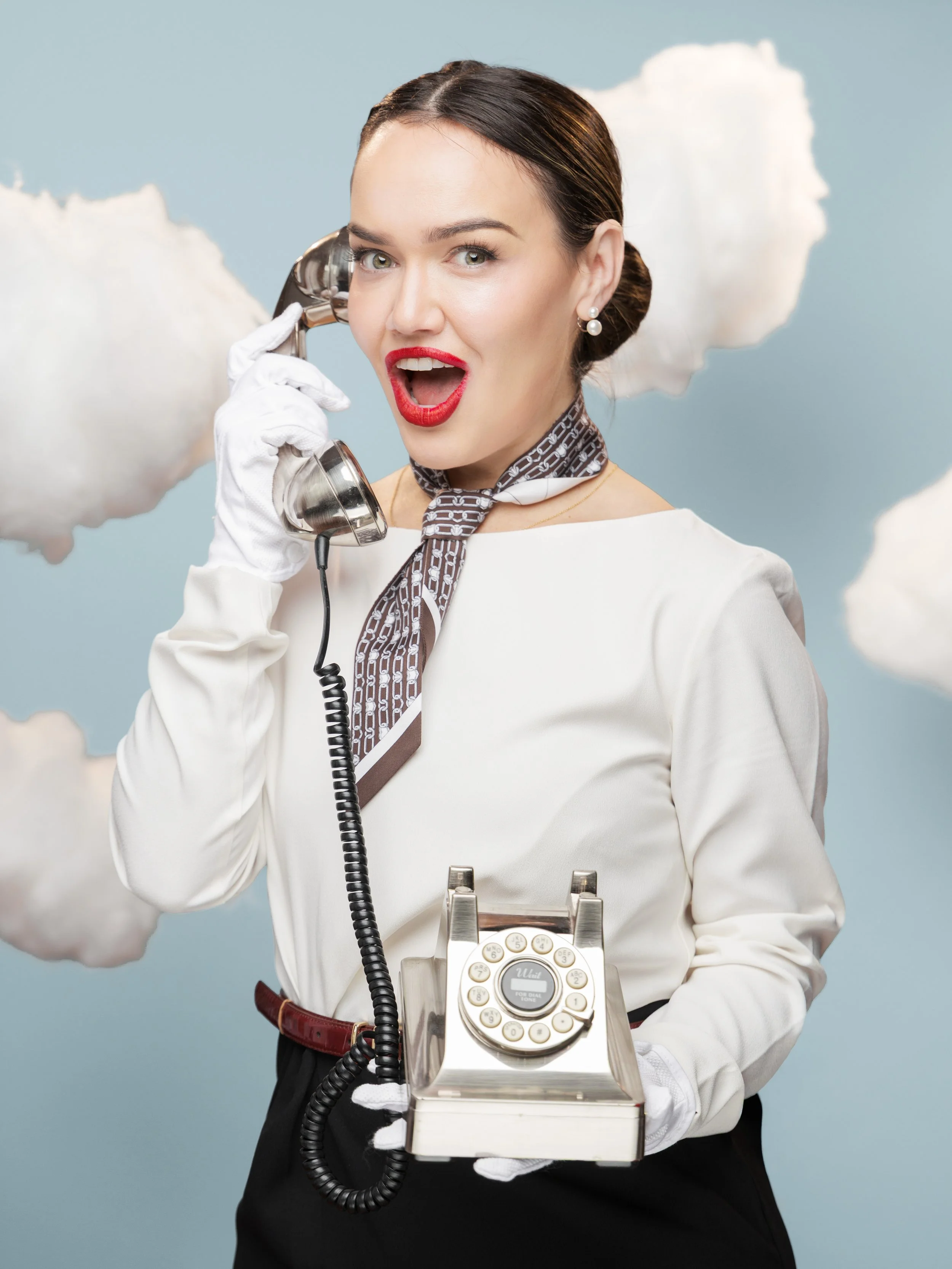 A woman in vintage attire with red lipstick and pearl earrings is holding a rotary phone to her ear with a surprised expression. She is also holding the phone's base in her other hand, and the background features clouds against a light blue sky.