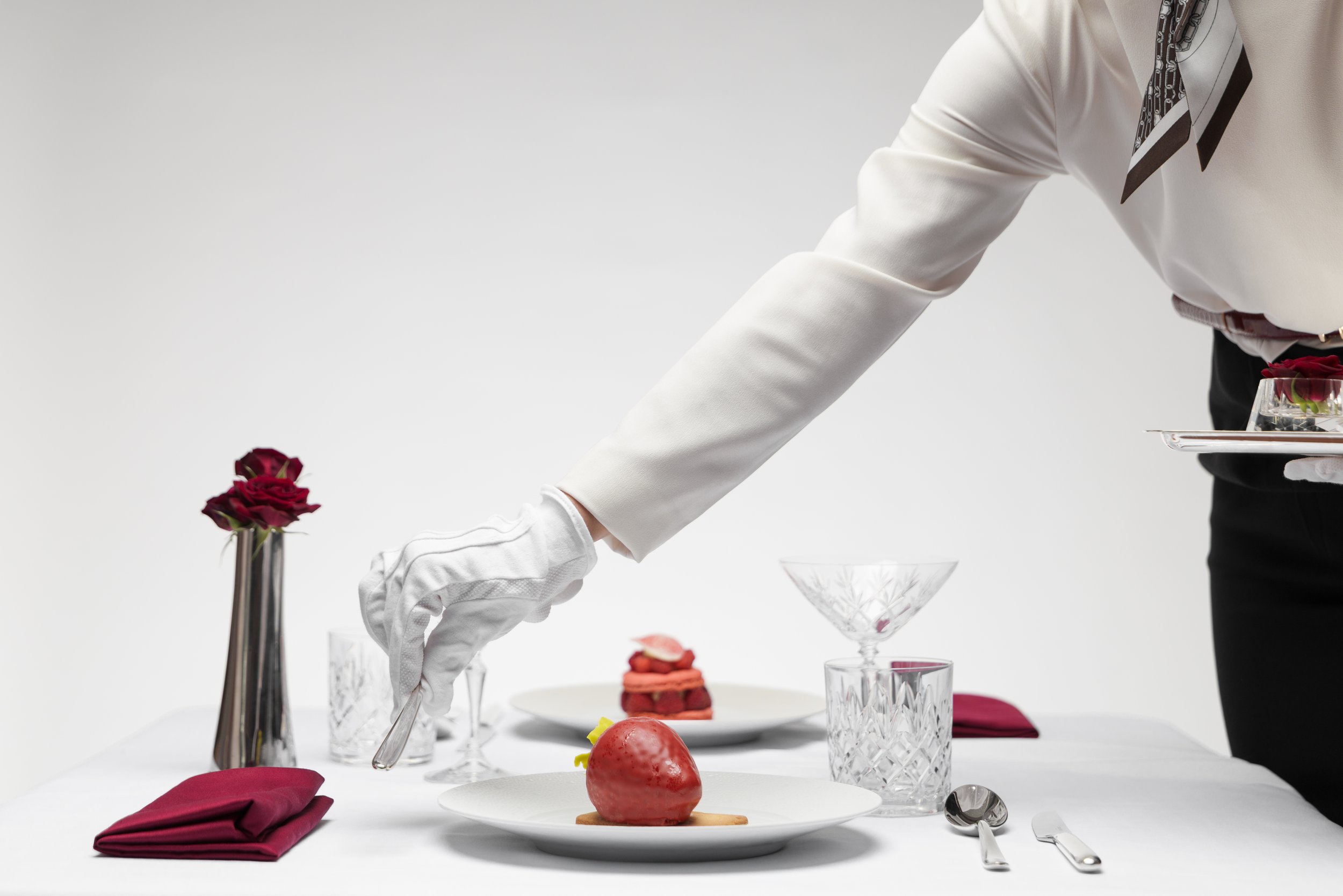 Person in white jacket and black pants setting a luxurious dining table with elegant glassware, red roses in a tall silver vase, and plates of gourmet food including a strawberry dessert and a layered pastry.