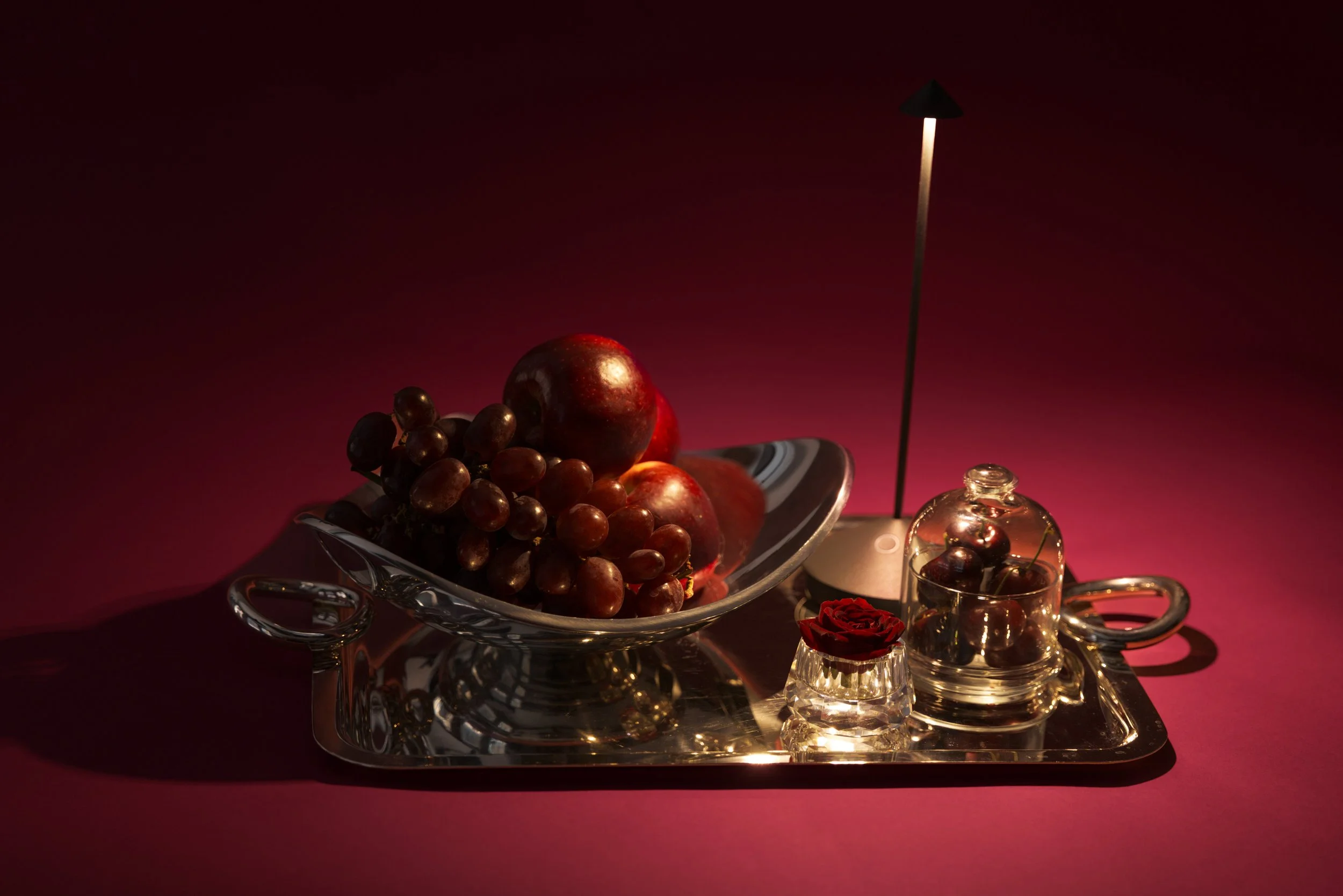 Still life of grapes and apples on a silver tray, with a small glass dome, a red rose in a glass container, and a tall candle, all on a silver tray against a dark pink background.
